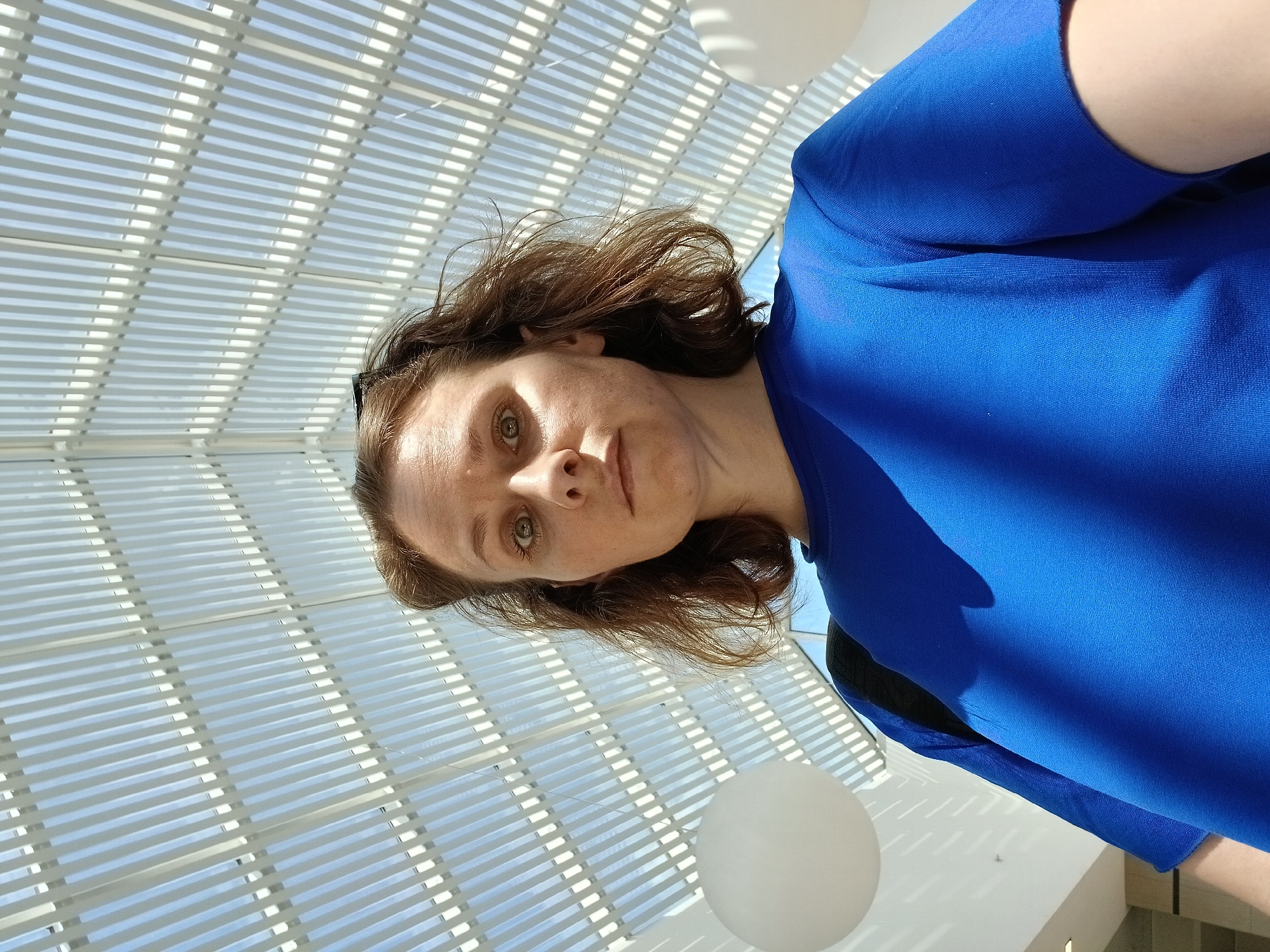 An indoor portrait shows a woman with medium-length brown hair and light skin looking directly at the camera. She is wearing a bright blue t-shirt. Her eyes are wide, and her mouth is slightly open. Behind her, a white, slatted ceiling casts shadows. Two large, spherical white lights hang in the background, one on each side of the frame. The setting appears to be a modern indoor space, possibly a public building or shopping center, with elements of architectural design visible in the ceiling structure and lighting. No specific location in Yekaterinburg, Russia is identifiable from the visual information present in the image.