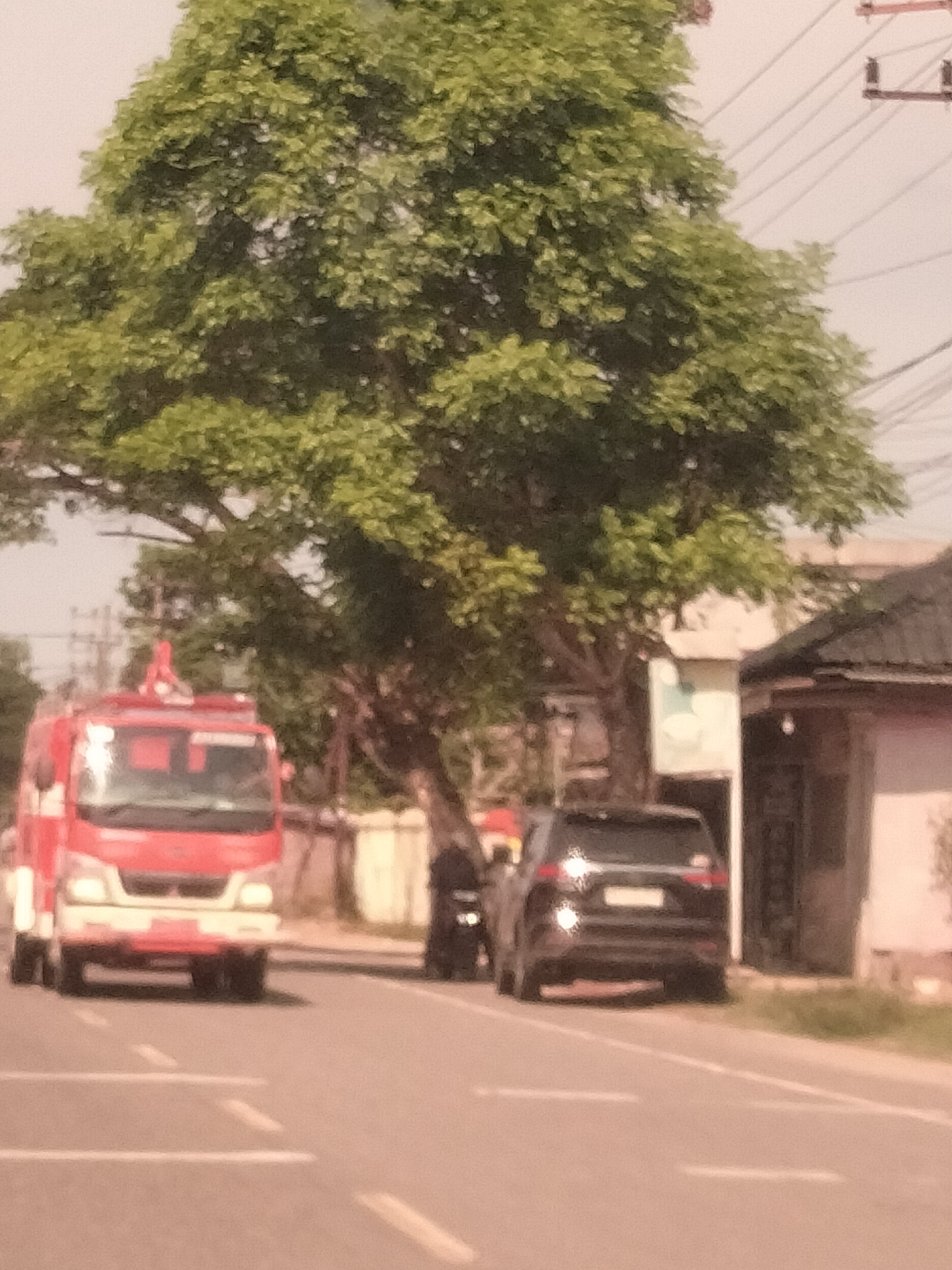 A slightly blurred image shows a red and white fire truck and a dark SUV parked on the side of a road in Meudangara, Indonesia. The fire truck is positioned on the left side of the frame, facing the viewer. A dark-colored SUV is parked to the right of the fire truck. A person is standing between the fire truck and SUV, partially obscured. The background includes a large tree with green foliage, power lines, and a building. The road has painted white lines.