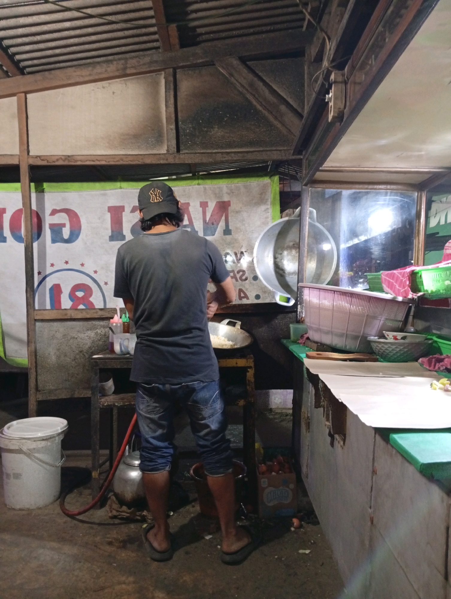 A person wearing a black baseball cap with a yellow logo and a dark t-shirt is standing with their back to the camera, facing a food stall in what appears to be an outdoor market at night. The person is cooking, with their hands in a pan on a stovetop. They are wearing blue denim shorts and flip-flops.
The food stall has a metal roof supported by wooden beams and a corrugated metal wall behind the person. A large banner with red and blue lettering, partially obscured, hangs on the wall. There are various cooking utensils and containers visible, including a large metal basin, plastic baskets, and bowls. To the left of the person, there is a white plastic bucket and a silver kettle. To the right, a counter is covered with paper, and various items are placed on it, including food ingredients and cleaning supplies.
The ground is made of dirt and concrete. The overall impression is of a humble, active food preparation area within a bustling night market. The location is identified as the city of Caringin, Indonesia.