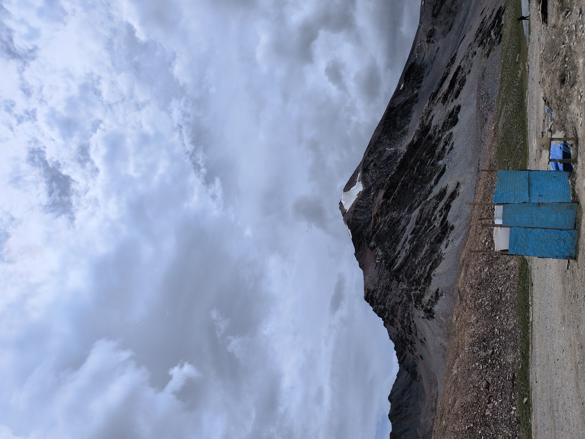 The image depicts a mountainous landscape under a cloudy sky. In the foreground, a rudimentary blue structure, possibly a portable toilet or shelter, is visible. Beyond the structure, a vast expanse of rocky terrain leads to a large, imposing mountain with patches of snow on its summit. The mountain's slopes are characterized by rugged, layered rock formations. The sky is filled with heavy, gray clouds, suggesting overcast or impending weather. A single human figure can be seen walking on the right side of the image, near the base of the mountain. The ground in the foreground is a mix of gravel and dirt. The overall scene conveys a remote, rugged, and possibly desolate environment. The location is identified as Killingsarai, India.