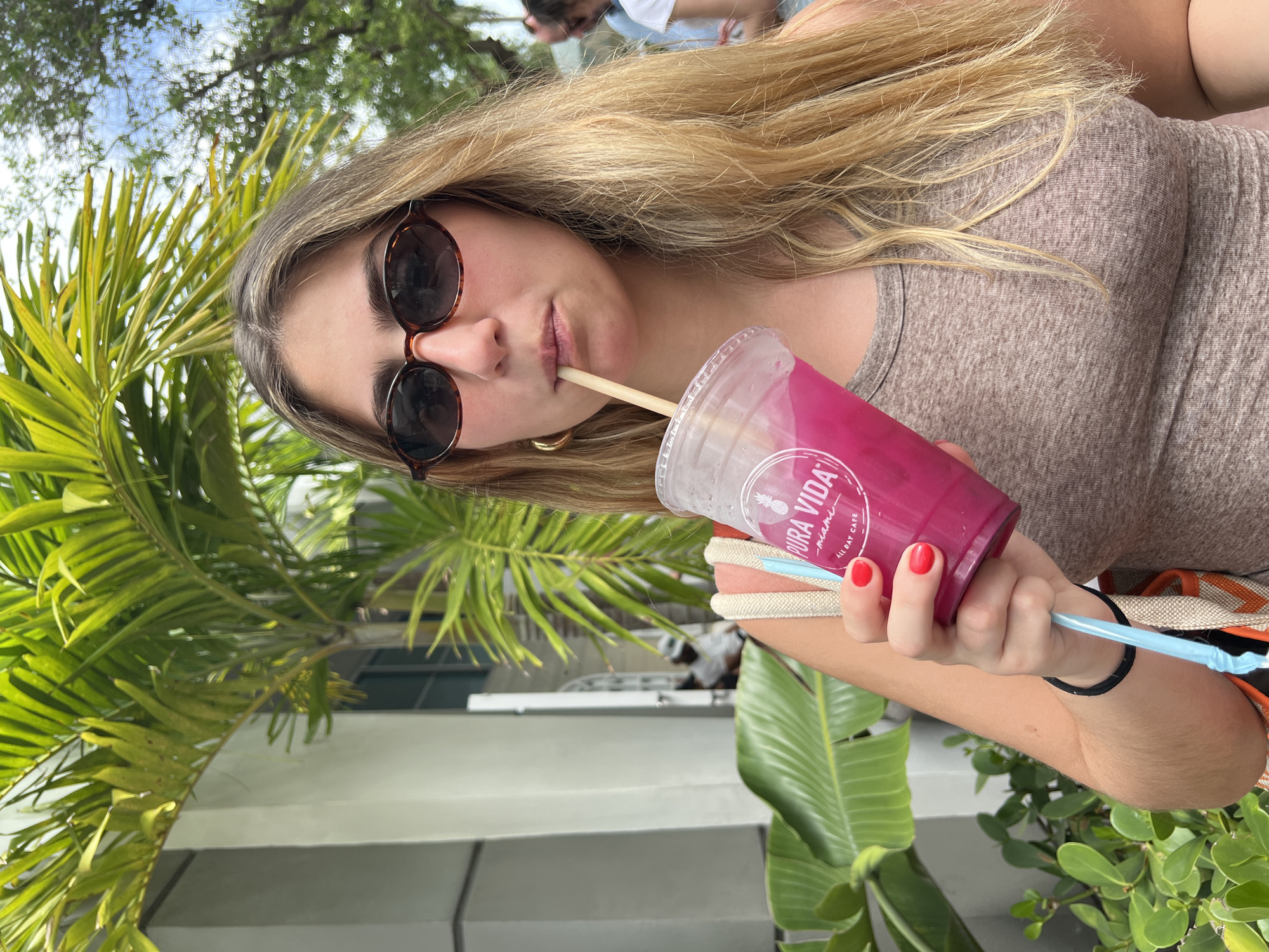 A young woman wearing sunglasses and a brown shirt drinks from a pink beverage cup labeled "Pura Vida, Miami, All Day Cafe." She uses a brown straw and holds a blue straw in her hand. The background includes lush green foliage, potentially palm trees and other tropical plants, suggesting an outdoor setting. A white structure is partially visible. The location is Miami Beach, United States of America. Other people are blurry in the background. The woman's nails are painted red.