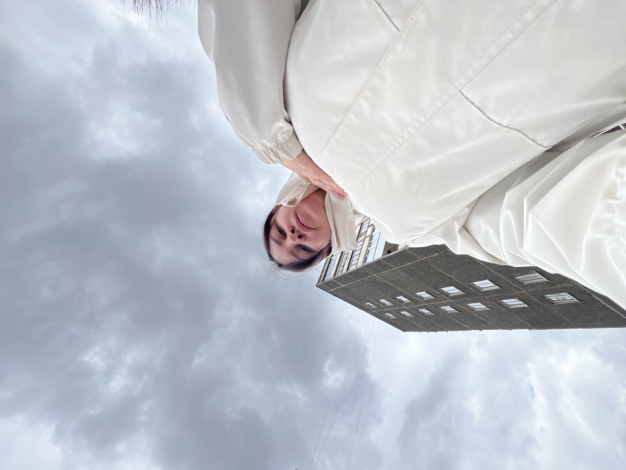 A low-angle, upside-down view shows a woman in a light-colored jacket standing outdoors, likely in Perm, Russia. The sky is overcast. A building with multiple windows is visible in the background to the right. The woman's face is positioned towards the center, and she appears to be holding her hand near her neck. There are power lines stretched across the sky.