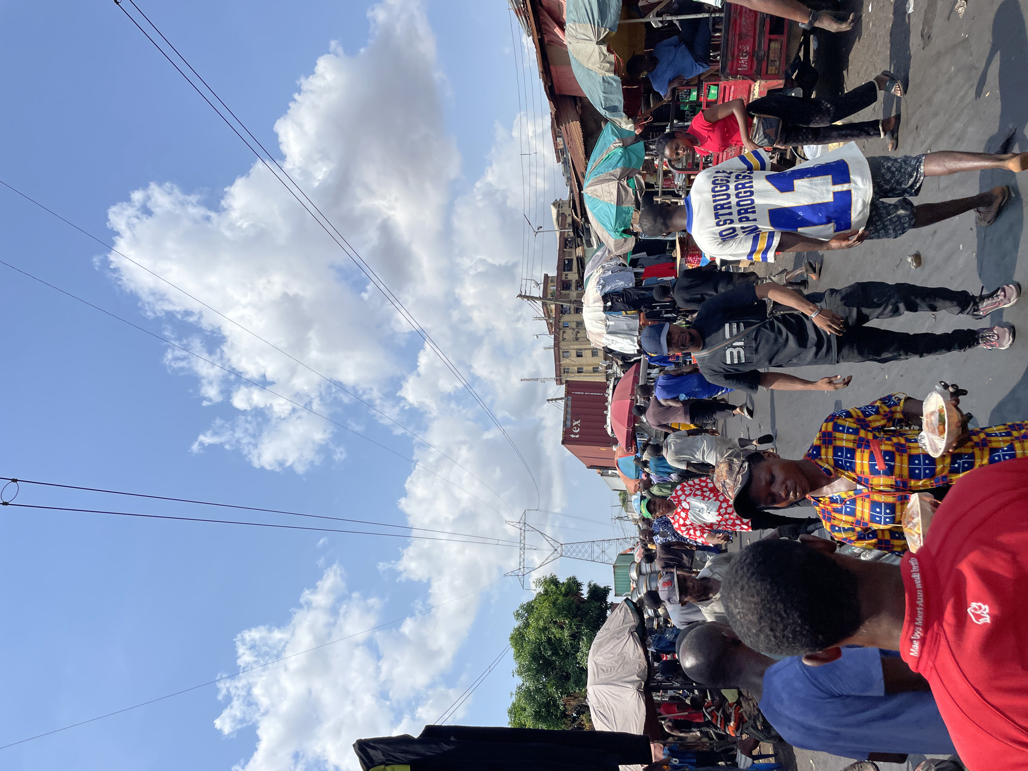 This media file captures a vibrant and busy street market scene in Onitsha, Nigeria, under a bright, sunny sky with scattered white clouds. The image, taken in daylight (likely midday or early afternoon given the strong lighting and shadows), shows a densely populated area filled with people, market stalls, and various urban infrastructure.
Numerous individuals are visible throughout the frame, engaged in activities typical of a bustling market. Many are walking, standing, or gathered in small groups. Some appear to be vendors, with makeshift stalls and goods partially visible, while others are shoppers or passersby. In the foreground, the back of a person wearing a red polo shirt is seen, with text on the collar that appears to read "Mae bye Mori Agu wadi bren." Next to them, a person in a blue shirt and another in a yellow, blue, and red patterned shirt are visible, the latter holding a clear plastic container, possibly containing food.
Further into the scene, a man in a black shirt and cap carries a shoulder bag with "B3" printed on it. A prominent figure is a man wearing a white and blue athletic jersey with the text "NO STRUGGLE NO PROGRESS" and the number "42" on the back. A woman in a red top and black trousers stands nearby.
The setting features various market structures, some with corrugated iron roofs, lining the street. Several open umbrellas, in shades of blue, white, and green, provide shade over stalls or individuals. Overhead, multiple power lines crisscross the blue sky. In the mid-ground, a large electricity transmission tower (pylon) stands tall, a notable landmark. Buildings, some multi-storied, are seen behind the market activity. A red shipping container, partially visible on a structure in the upper right, has "tex" printed on it. The ground appears to be a paved or unpaved street surface. The overall atmosphere is one of active commerce and daily life in a crowded urban environment.