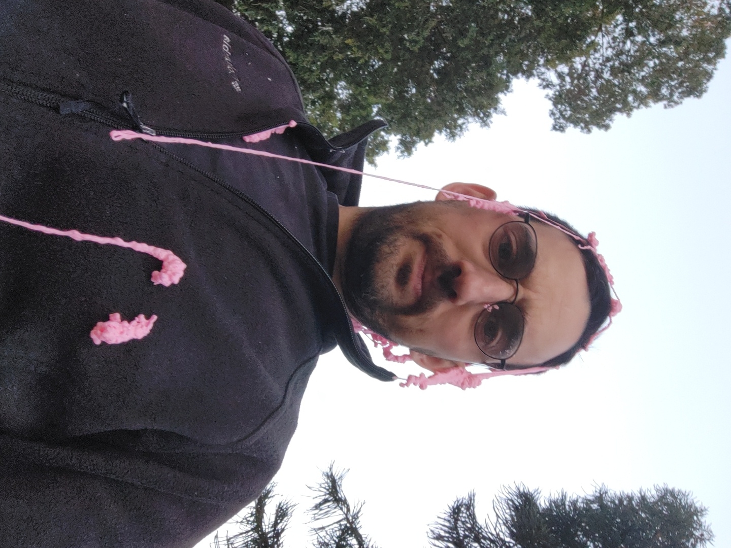 The image, taken in Tel-Aviv, Israel, shows a man outdoors, looking directly at the camera from a low angle, giving the impression of a selfie or a shot taken from below. The photo is oriented with the man's body appearing vertical and his head horizontal.
The man has a beard and is wearing round glasses. He has light pink, string-like material, possibly chewing gum or cotton candy, stuck to his hair, face, particularly on his right ear, and around his forehead. Similar pink material is also visible on the black fleece jacket he is wearing, appearing as several distinct strands and clumps on the chest area.
The black fleece jacket features a zippered front, with a visible zipper pull on the right side. Partially visible text on the jacket includes "BLOKHAUS" and the "adidas" logo.
In the background, against a bright, overcast sky, trees are visible. The upper portion of the image shows the canopy of a deciduous tree, while the lower part shows darker, sparser tree branches, possibly conifers, suggesting a park or outdoor setting. The overall lighting indicates it is daytime. The man's expression is neutral, possibly hinting at amusement or resignation due due to the pink substance.