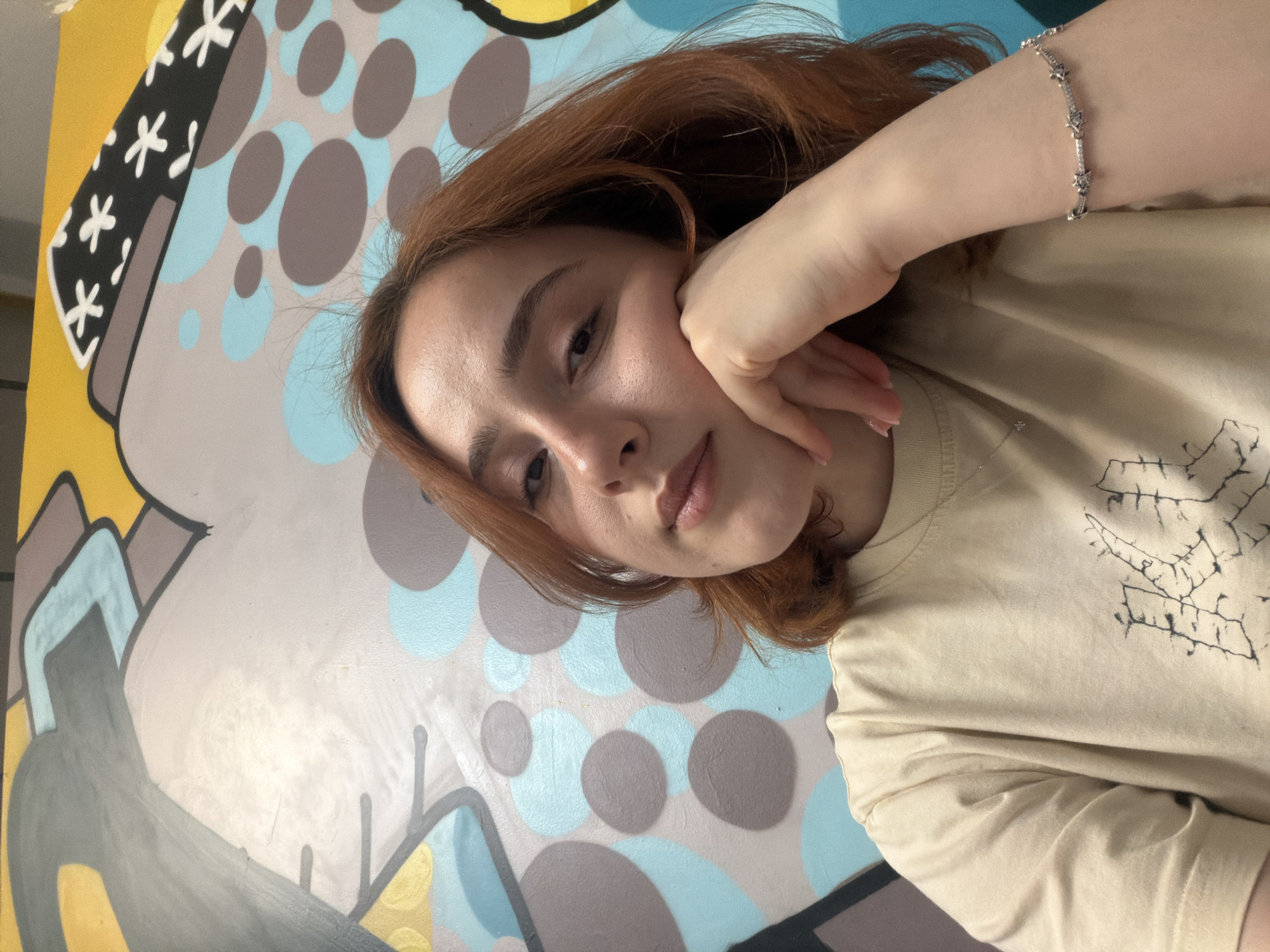 A young woman with auburn hair, wearing a beige t-shirt and a delicate bracelet, is captured in a selfie. Her left hand rests on her cheek, and she looks directly at the camera with a neutral expression. She is positioned in front of a vibrantly colored mural that features abstract shapes, circles in shades of blue and grey, and geometric patterns in yellow and grey. The mural appears to be on a wall, likely in an urban setting. There is no indication that this location is in Strasbourg, France.