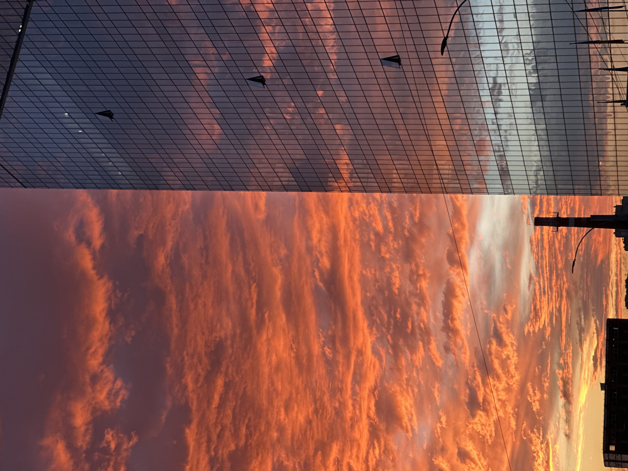 The image captures a dramatic sunset over the city of Chișinău, Moldova. The sky is filled with vibrant hues of orange and red, painting a breathtaking panorama of clouds. A modern skyscraper with a glass facade dominates the right side of the frame, its windows reflecting the fiery sky and the surrounding cityscape. On the left, a lower industrial-looking building, possibly a factory or a power plant, is silhouetted against the colorful sky. Streetlights and power lines are visible, hinting at the urban environment. The overall impression is one of urban beauty juxtaposed with the raw power of nature's spectacle.