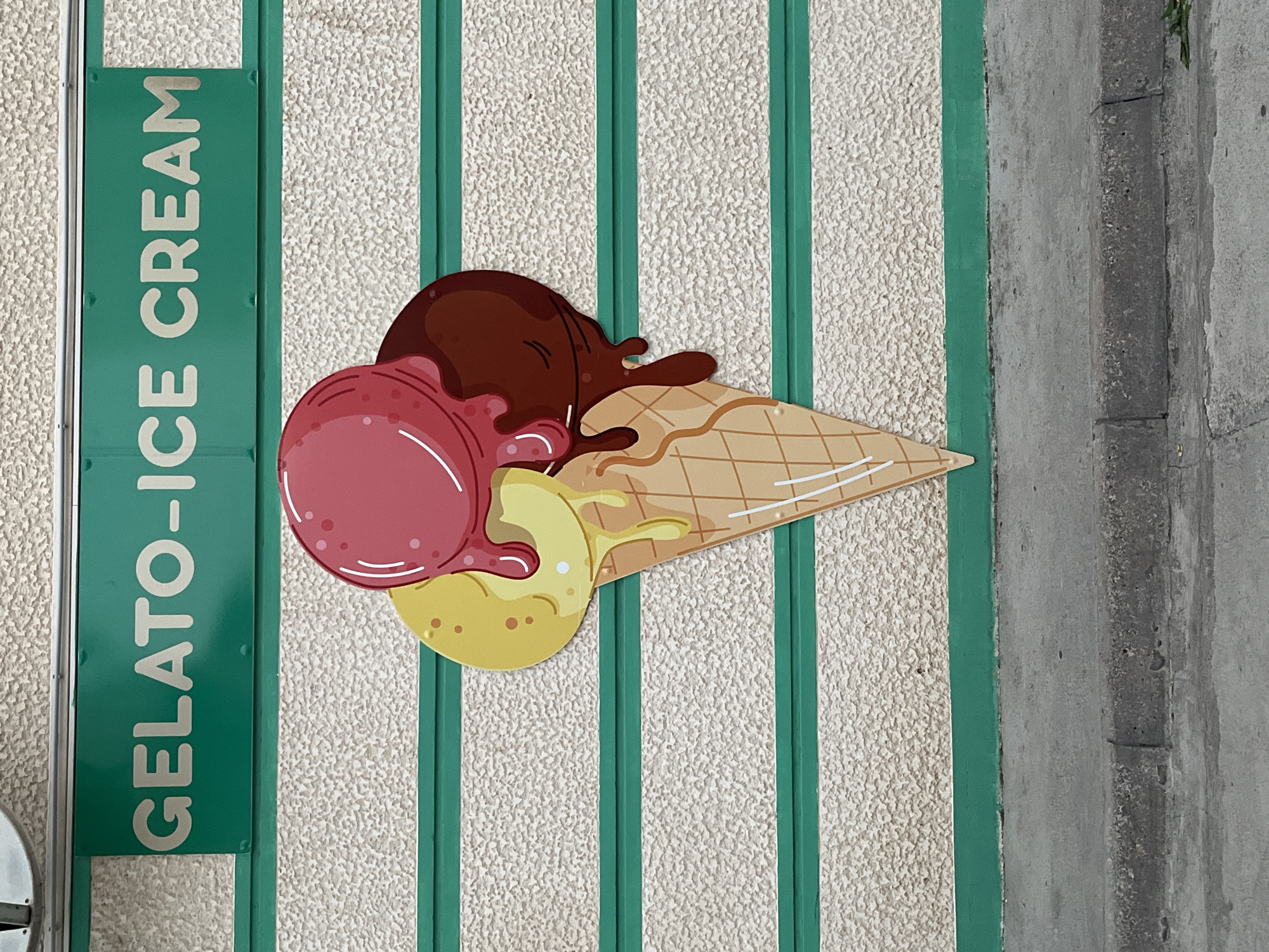 The image shows a flat, cut-out depiction of a three-scoop ice cream cone with melting drips, made of colored material. The ice cream scoops are pink, yellow, and dark brown. The cone is light brown with a waffle pattern. Above the ice cream is a horizontal green sign with white lettering that reads "GELATO - ICE CREAM". The sign and the ice cream are mounted on a textured wall with horizontal green stripes. Below the wall is a concrete sidewalk and curb. The scene is outdoors, with natural light. This appears to be a decorative sign for a gelato or ice cream shop. It is not possible to determine the specific location in Budva, Montenegro based on the image alone.