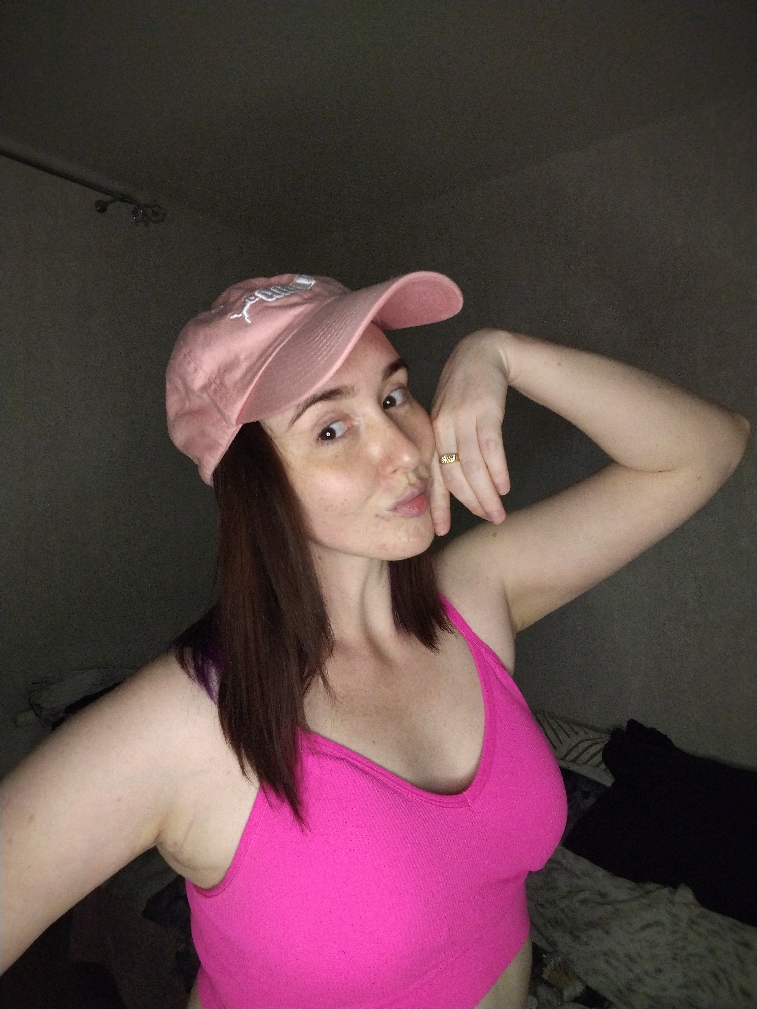 A woman is posing in a dimly lit room. She is wearing a pink baseball cap and a pink tank top. Her dark hair is shoulder-length. Her right arm is raised, with her hand near her face. A gold ring is visible on her finger. The background is dark and indistinct, showing a ceiling fixture in the upper left corner and bedclothes. No specific location in Izhevsk, Russia, can be determined from the image.