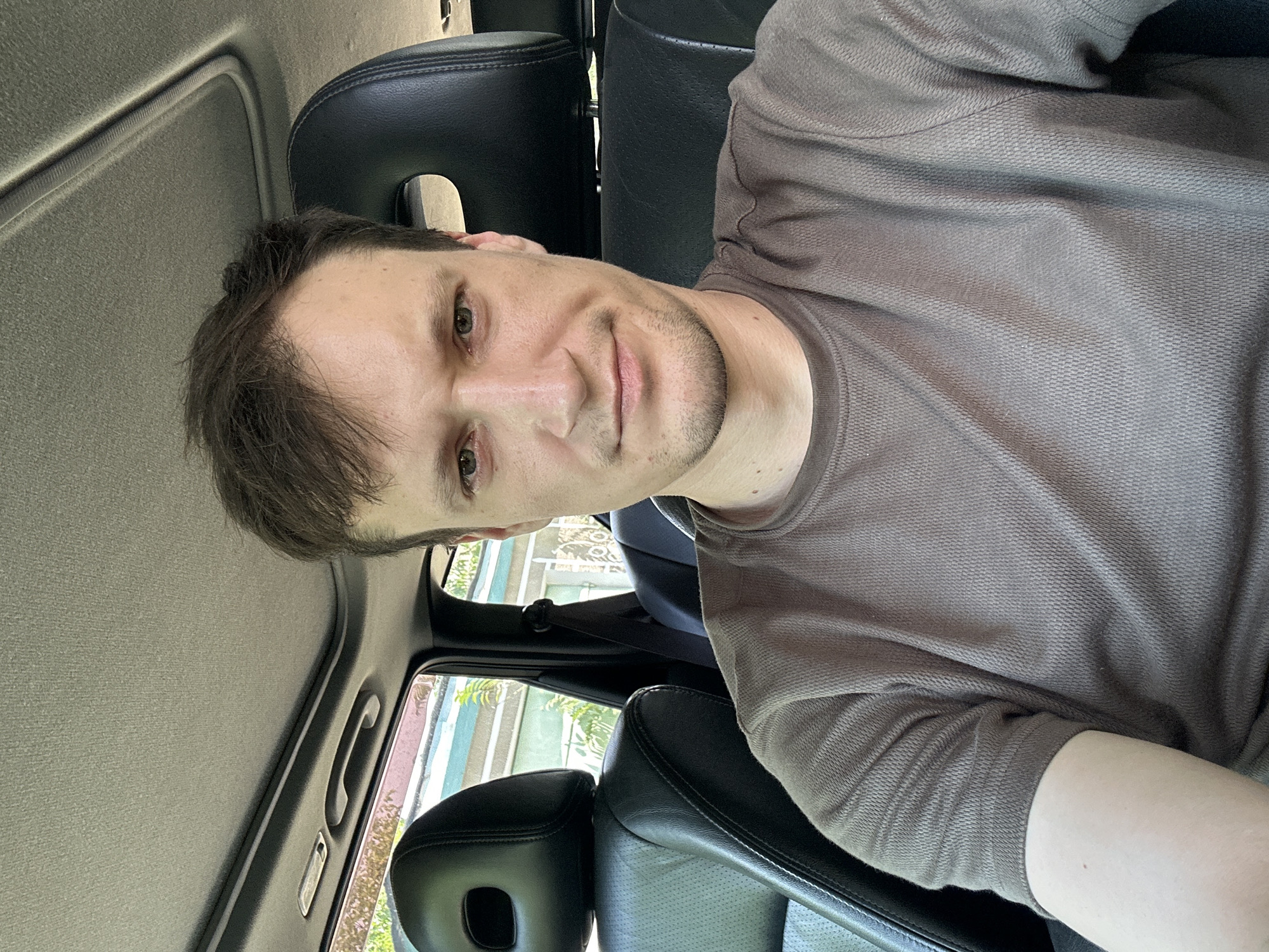A man with short brown hair and light green eyes is seated in the driver's seat of a car. He is wearing a short-sleeved, brown t-shirt. The interior of the car is visible, including black leather seats, the car's ceiling with a sunroof, and a seatbelt. Outside the car, through the windows, greenery and a building facade are discernible. The image appears to be a selfie taken inside the car. The location is not discernible from the image.