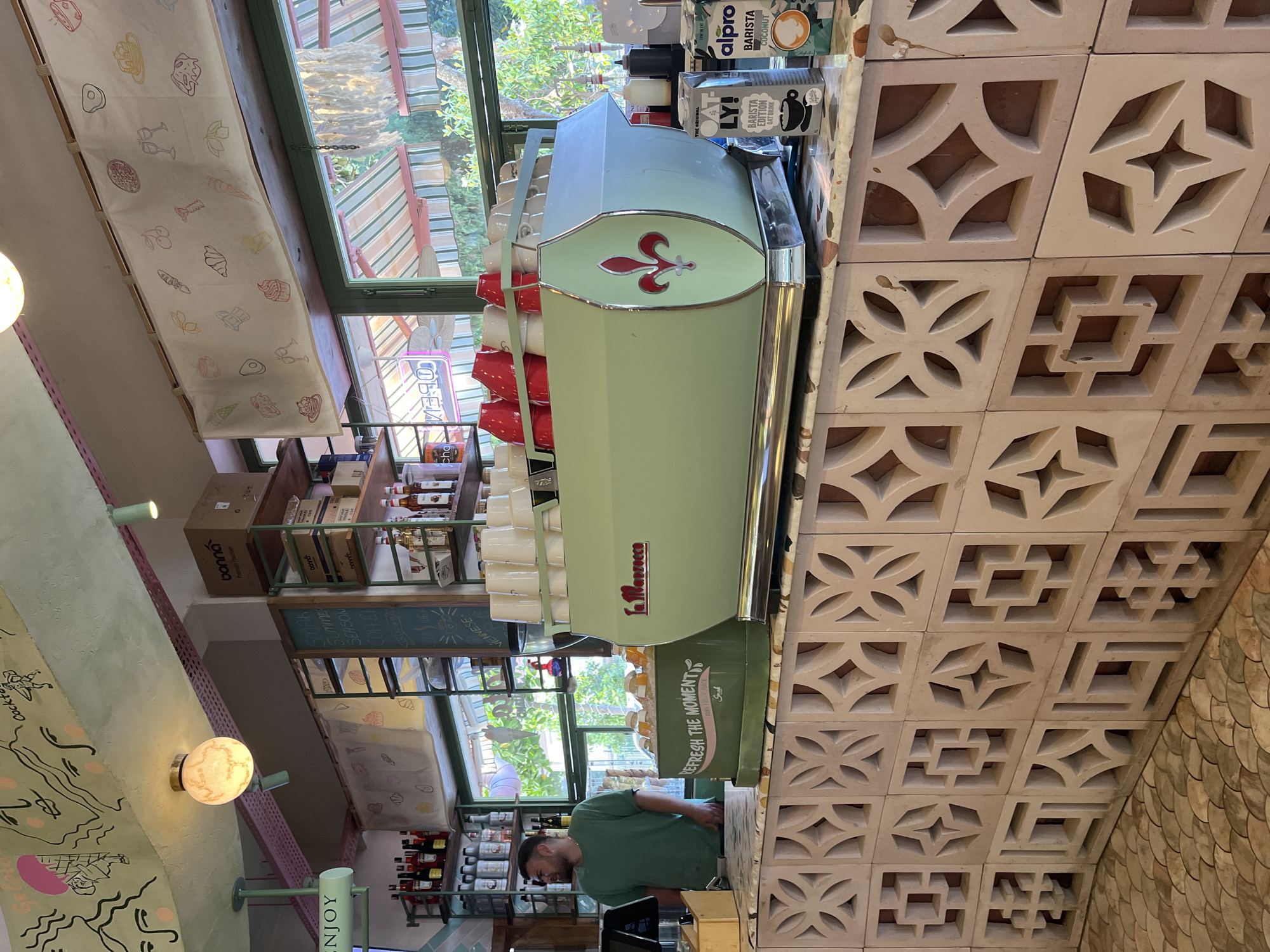 A vertically oriented medium indoor shot shows the interior of the "SMAK" cafe in Alanya, Turkey. A man wearing a green t-shirt is visible behind a light-green La Marzocco coffee machine, which has multiple cups stacked on top. Above the machine, brands of milk alternatives are displayed, including Alpro and Ly! Barista Editions. To the right, a wall is covered in decorative geometric beige tiles. Behind the coffee machine, a window allows natural light into the cafe. Visible through the window are outdoor elements, including greenery. A shelf unit holding bottles and containers is situated near the window. A light fixture with a marbled glass globe is attached to a painted wall with artistic drawings and the word "ENJOY" printed on a pole in green and white letters. A curtain with food-related sketches hangs near the top of the window. Additional branding and menu boards can be seen within the cafe.