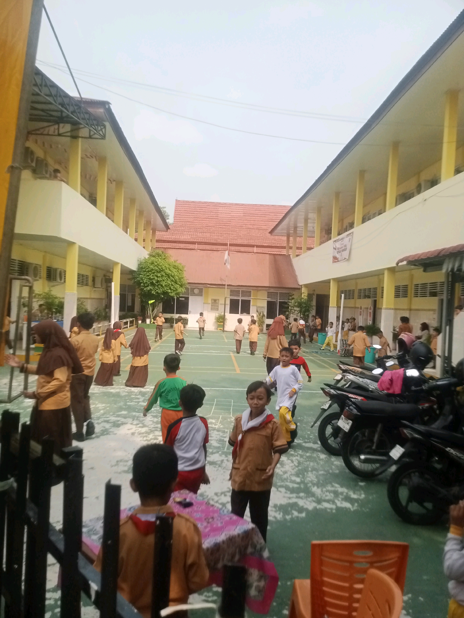 The image depicts a schoolyard in Pekanbaru, Indonesia, on an overcast day. In the foreground, several children are visible. One child in a brown scout uniform stands facing the camera, with a patterned cloth-covered table and an orange chair to their right. To the left, a dark metal fence is prominent. In the midground, a group of children in brown uniforms, some wearing hijabs, are walking on a green court. Other children in similar uniforms are seen scattered across the court, with some engaged in activities. In the background, a two-story yellow building with white columns lines one side of the court, while another building with a red tiled roof is visible on the opposite side. Several motorcycles are parked along the side of the court, to the right of the frame. The overall scene suggests a school event or recess.