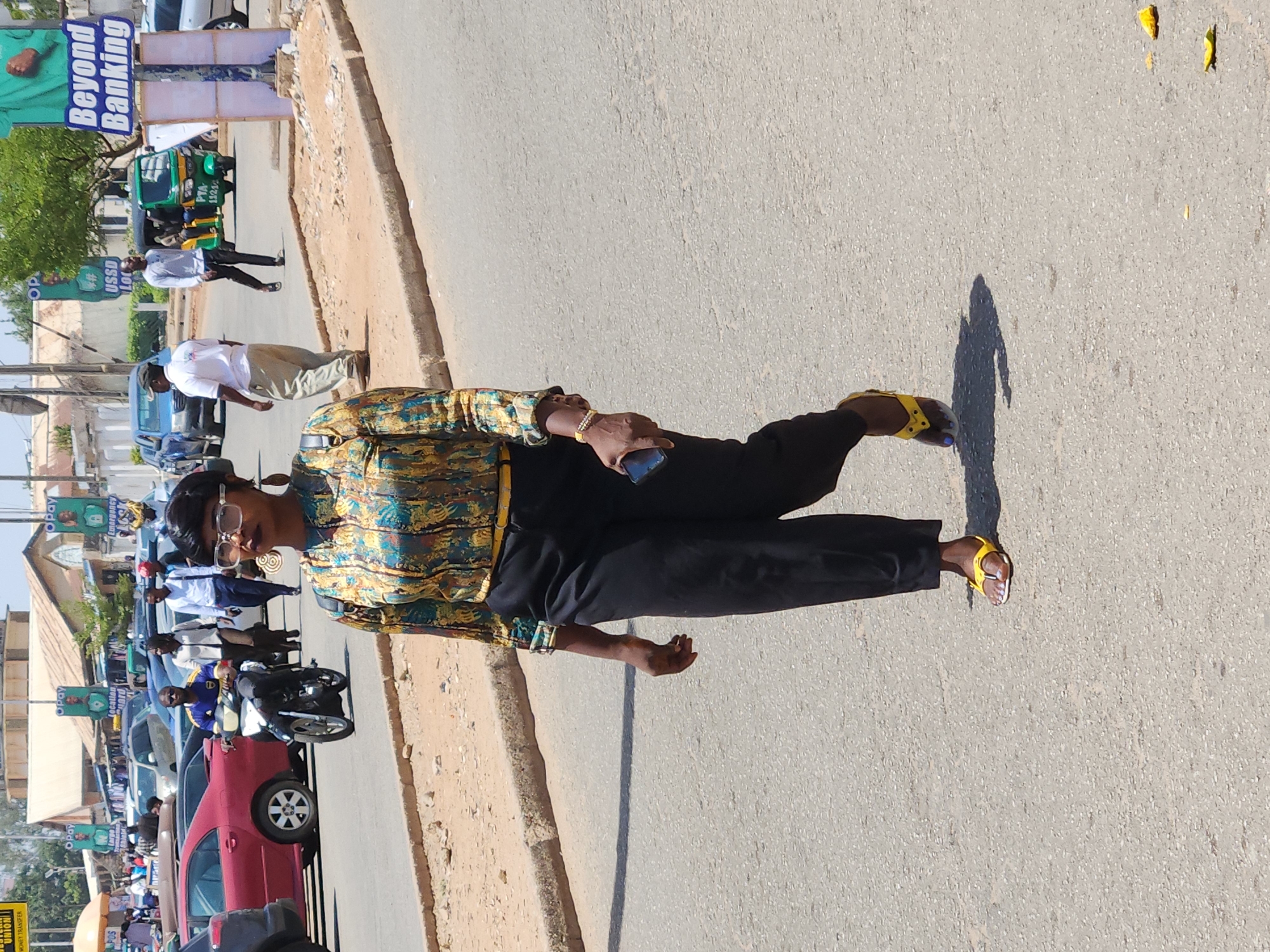 The image captures a woman standing on the side of a road in an unidentified city. She is dressed in a long-sleeved shirt with a gold, blue, and green pattern, paired with black pants and yellow sandals. She holds a smartphone in her right hand and wears glasses and earrings. In the background, there's a street lined with cars, motorcycles, and people. Signage for "Beyond Banking," "Opay," and "MONEY TRANSFER" is also visible, as is a three-wheeled vehicle. The setting suggests a bustling urban environment.
