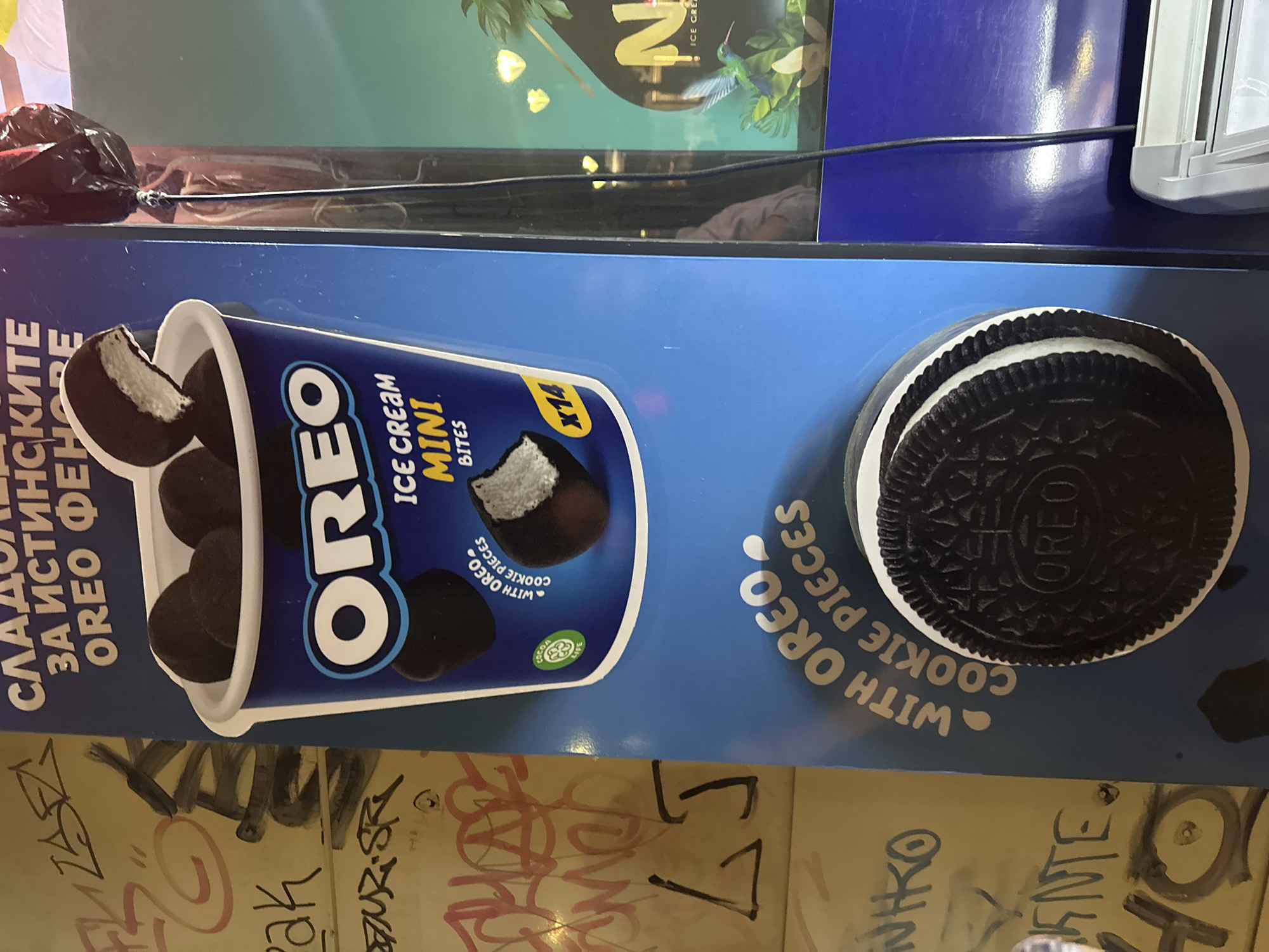 A blue vertical display advertises Oreo Ice Cream Mini Bites. The top of the display features text in Cyrillic, followed by an image of a cup filled with dark, round ice cream bites with white creamy centers. The Oreo logo is prominently displayed in white text on a dark blue background. Below the cup, smaller text reads "ICE CREAM MINI BITES". To the right of this section, a yellow circle with "x14" is visible. Further down, text in white, curved lettering states "WITH OREO. COOKIE PIECES". A large, circular Oreo cookie is depicted at the bottom of the display, showing its distinct pattern and creamy white filling. The background on the left side of the display is covered in graffiti, with various tags and scribbles in different colors, including red, black, and yellow. The background on the right is a blurred image of what appears to be a tropical scene with palm trees and a hummingbird, with some text and logos visible. The display is illuminated, and a thin black cable hangs vertically on the right side of the image.
