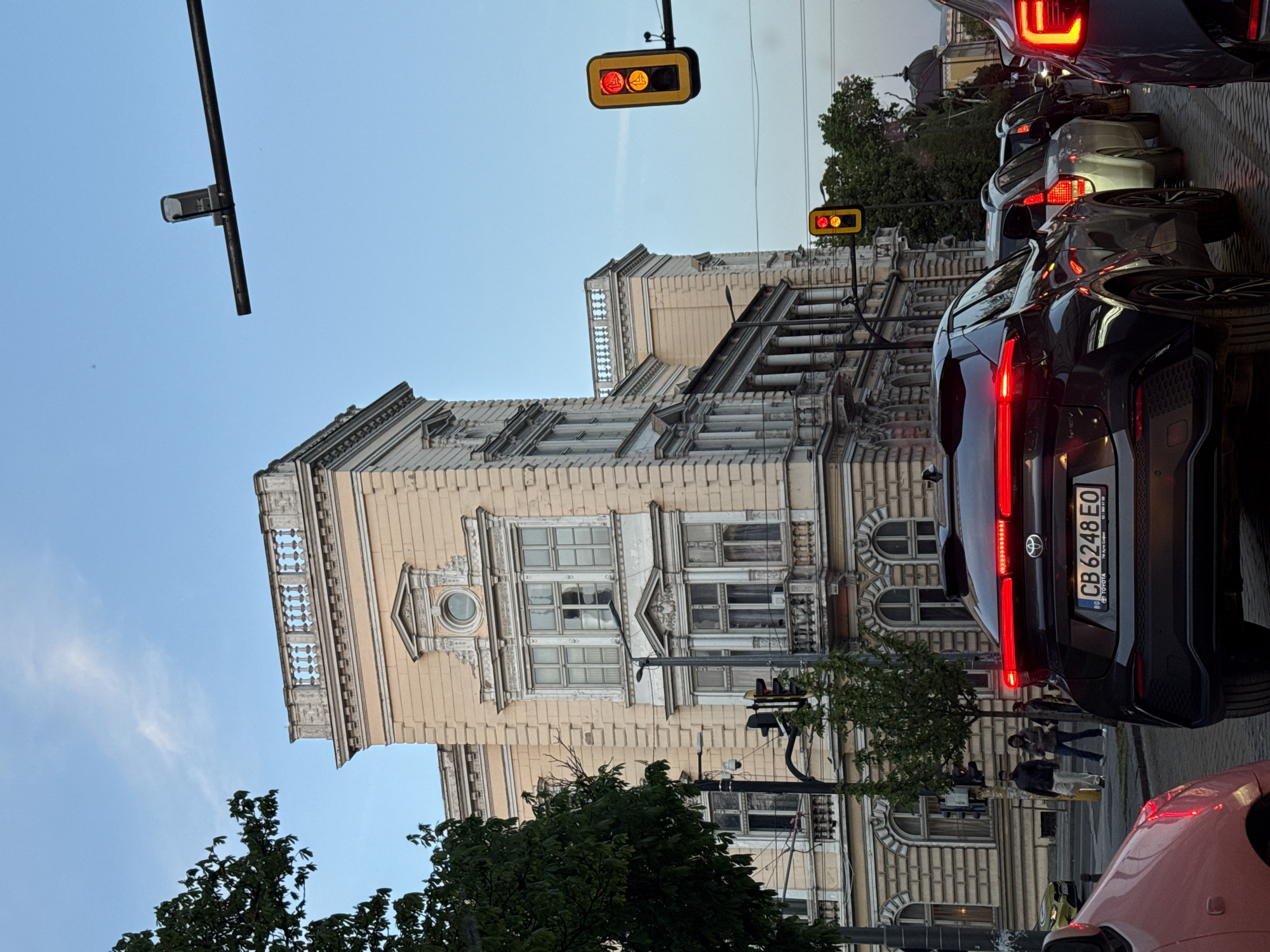 The image depicts a busy city street scene at dusk or early evening. Several cars are stopped in traffic, with their red brake lights illuminated. A prominent black Toyota C-HR SUV is visible in the foreground, displaying a Romanian license plate that reads "CB 6248 EO". Below the Toyota branding, the text "TOYOTA C-HR" and "RO" are visible.
To the left of the street, a grand, multi-story building with ornate classical architecture stands tall. Its pale, possibly cream or light yellow, facade is adorned with intricate decorative elements, including balconies or balustrades, and detailed window frames. The architecture suggests a historical or significant structure.
Above the street, two overhead traffic lights are visible. Both lights show illuminated red and yellow signals, with symbols that appear to be walking figures, indicating pedestrian traffic. Patches of green foliage from trees are interspersed throughout the urban environment, and a few pedestrians can be seen walking on the sidewalk next to the building. The sky above is a clear, light blue, suggesting a transition from day to night. The scene captures the typical atmosphere of an active urban center during the late hours of the day.