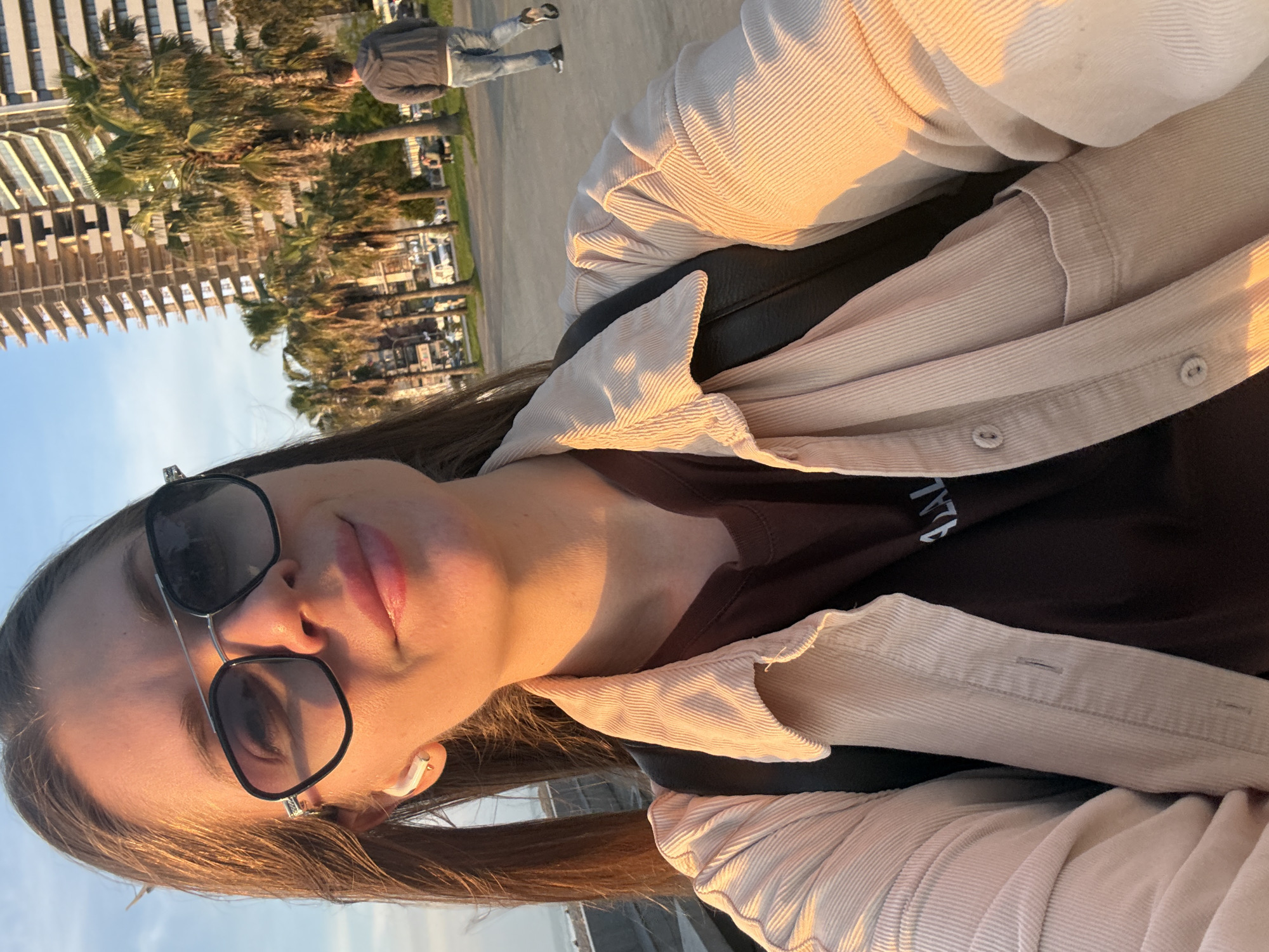 A young woman wearing sunglasses, a brown t-shirt, and a light corduroy jacket is standing outside. She is also wearing a white wireless earpiece in her left ear. The background features palm trees, modern buildings, and a sidewalk where a man is walking. The photo appears to be taken at sunset, indicated by the warm lighting. Based on the architecture and palm trees, the location is likely in the city of Batumi, Georgia.