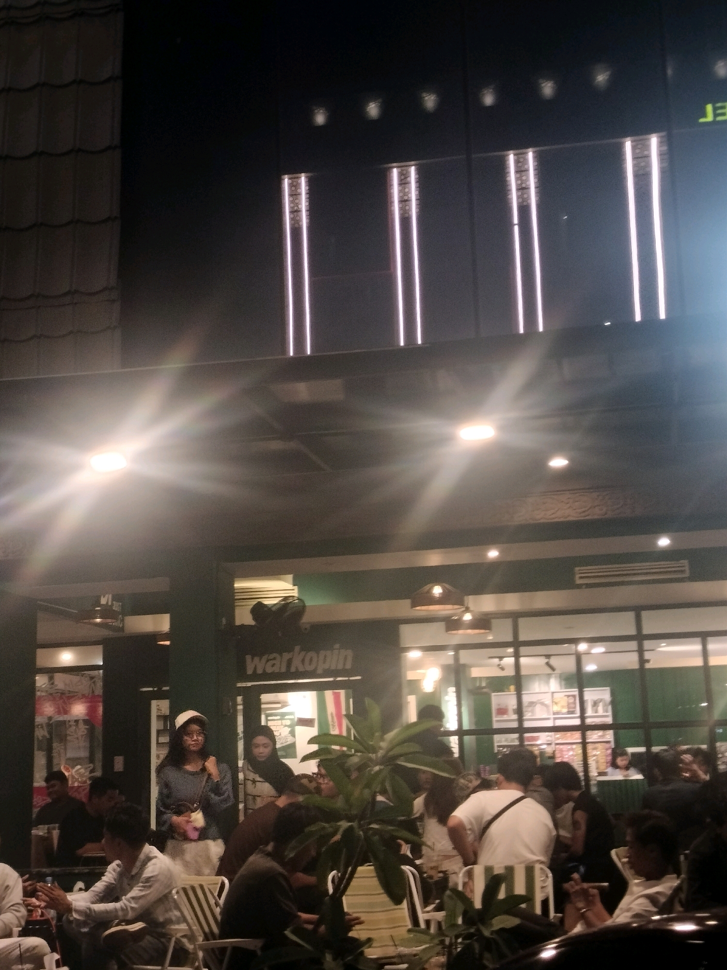 The image depicts an outdoor seating area of a "Warkop" (warung kopi, or coffee shop) in Batam, Indonesia, at night. The setting is illuminated by overhead lights, creating a bright glare in the upper portion of the image, and the warm glow from inside the establishment. In the foreground, several people are seated at tables, some engaged in conversation, while others appear to be smoking or looking at their phones. One individual in the mid-ground, wearing a blue long-sleeved shirt and a white cap, stands near the entrance of the establishment, facing the camera. Behind her, another woman is visible. The "Warkop" signage is prominently displayed. The establishment appears to be a popular gathering spot, with a lively atmosphere. The architecture of the building features large windows and a modern design. The presence of lush green plants adds a touch of nature to the urban environment. The overall scene suggests a casual and social evening at a local Indonesian coffee shop.