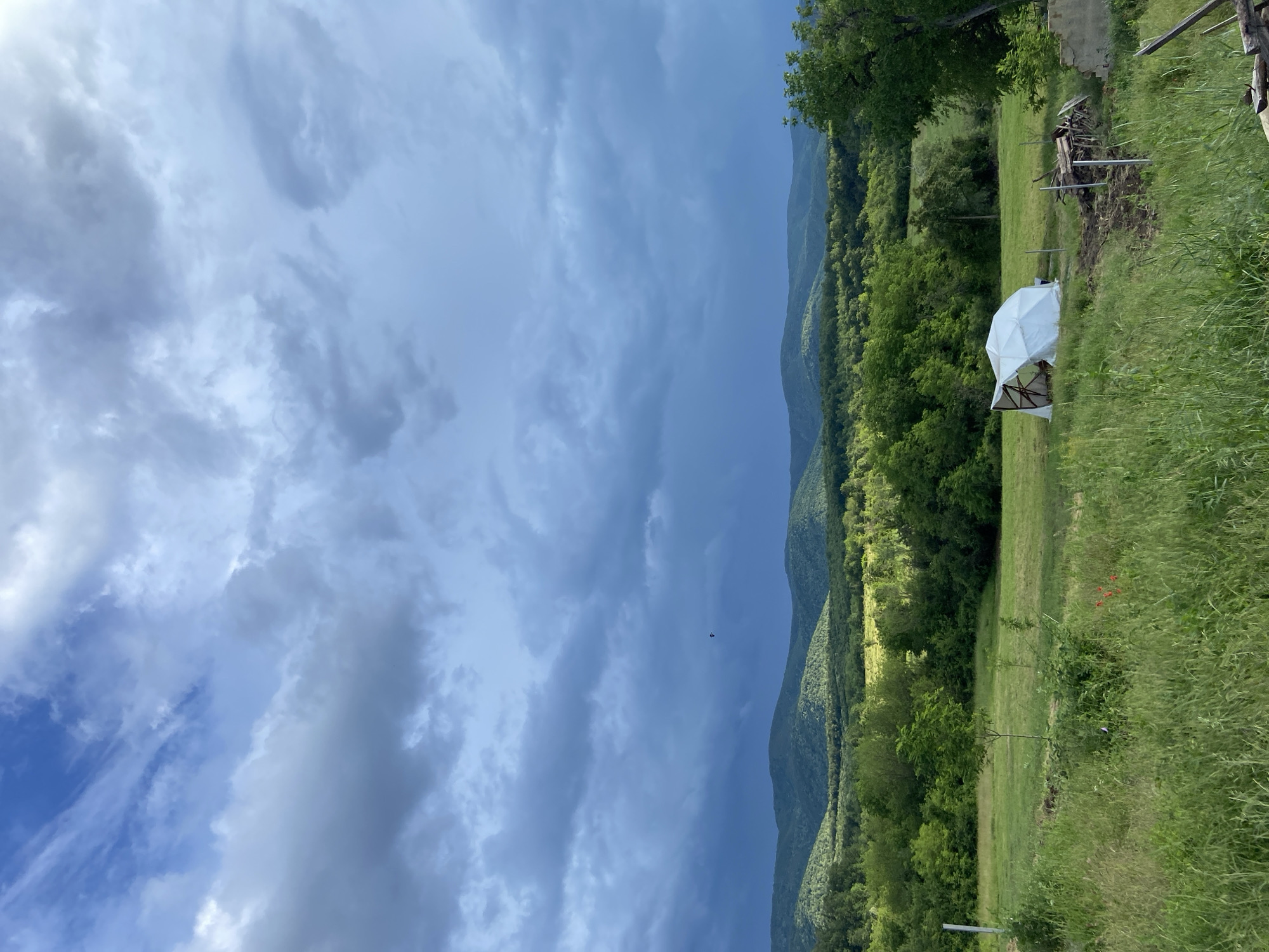 This media file presents a panoramic view of a lush, mountainous landscape under a dramatic cloudy sky, likely in Mindia, Bulgaria. The image is composed of three main horizontal sections.
The upper portion of the frame is dominated by a vast, cloudy sky. Dark grey, almost stormy-looking clouds are interspersed with lighter, brighter patches, suggesting diffused sunlight or breaks in the cloud cover. The overall impression is one of impending weather or the aftermath of a recent weather event.
Below the sky, a series of verdant, tree-covered mountains extend across the mid-ground. These hills rise and fall, creating a sense of depth and vastness. The slopes are densely forested with various shades of green, indicating abundant vegetation. The mountains in the distance appear slightly hazy and bluer, typical of atmospheric perspective.
The foreground consists of a large, grassy field with varying heights of green grass. On the right side of this field, a prominent white, temporary structure, possibly a geodesic dome tent or a large canopy, stands out against the natural backdrop. Its framework is discernible through the white material. To the left and slightly behind this structure, there's an area of disturbed earth with what appears to be discarded wood planks, brush, or construction debris. Further to the right, a dilapidated, light-colored stone or concrete structure is partially visible, hinting at an abandoned building or ruins. Some wooden posts or a simple fence line are also visible on the far right edge of the foreground. A few individual trees are scattered in the mid-ground, adding to the rural feel.
The time of day appears to be daytime, with soft, diffused light due to the heavy cloud cover. There is no visible text in the image. The scene evokes a sense of tranquil, rugged nature with subtle signs of human activity or presence.