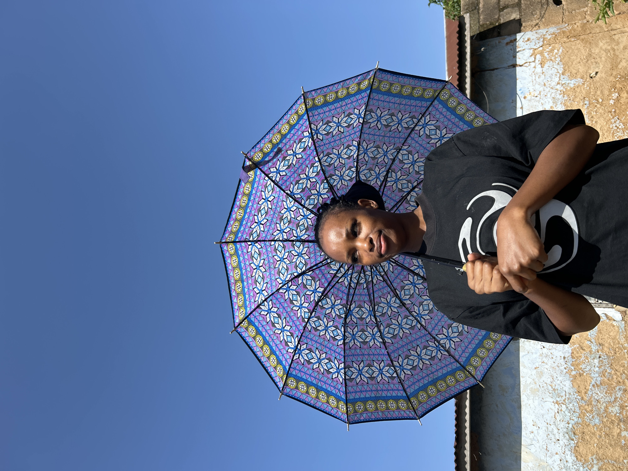 A young woman stands outdoors, holding an open umbrella with a purple, blue, and white floral pattern against a clear blue sky. She wears a black t-shirt with a white graphic. Behind her is a dilapidated wall with peeling paint, suggesting an urban or semi-urban setting, possibly in Jos, Nigeria, given the architectural style and vegetation. The sunlight casts a shadow on her face, and she looks slightly to the side with a neutral expression. The umbrella is positioned centrally, obscuring much of the background.