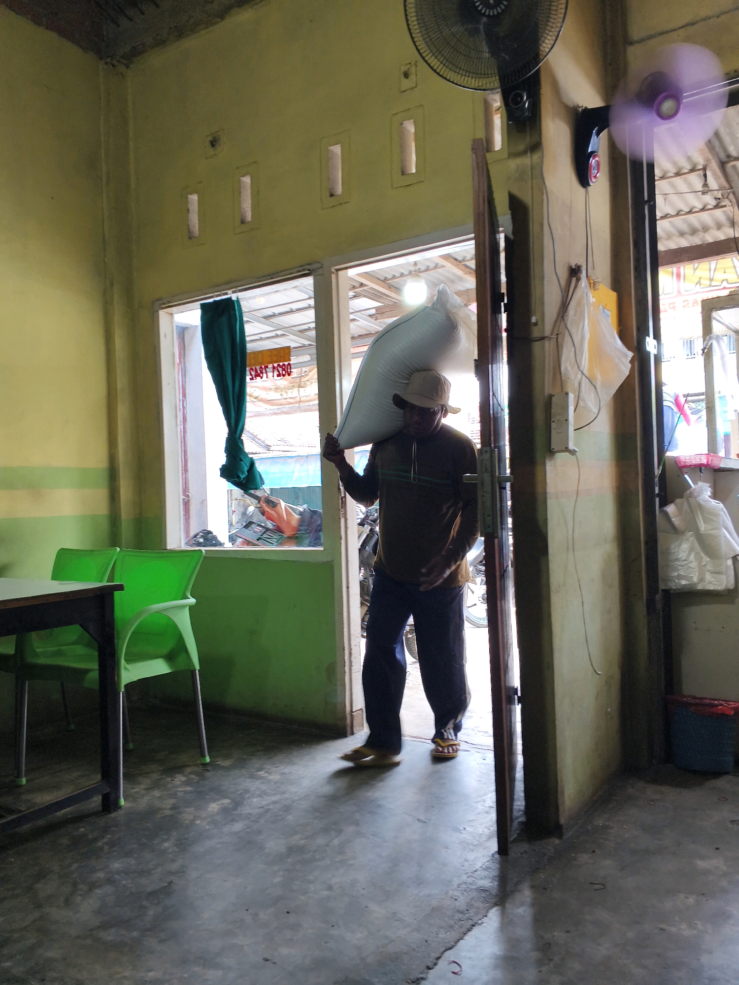 A man wearing a light-colored hat, a brown long-sleeved shirt with horizontal green stripes, and dark pants is carrying a large white sack over his right shoulder. He is walking through an open doorway into a room with pale yellow walls and a gray concrete floor. The doorway leads from an area with green walls, where two bright green plastic chairs and a dark table are visible. The room he is entering has a ceiling fan mounted high on the wall, and another fan with a purple blur is rotating on a different wall. Through a window in the green-walled area, a glimpse of a motorcycle and a green curtain can be seen. Bags and packaging materials are visible on shelves and hanging on the wall in the background. The overall impression is an indoor setting, possibly a shop or storage area, located in Sendang Retno, Indonesia.