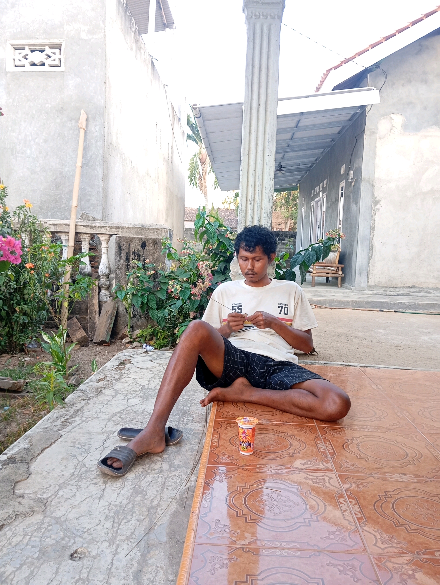A man is sitting on a tiled floor in front of a building in Leman Benawi, Indonesia. The man is wearing a light-colored t-shirt with graphics on it and dark shorts with a checkered pattern. He is holding something in his hands and looking down at it. There is a small cup with a picture on it to his right. The building behind him appears to be made of concrete and has a white overhang. There are plants and a bamboo pole visible in the background. The ground is a mix of concrete and tile.