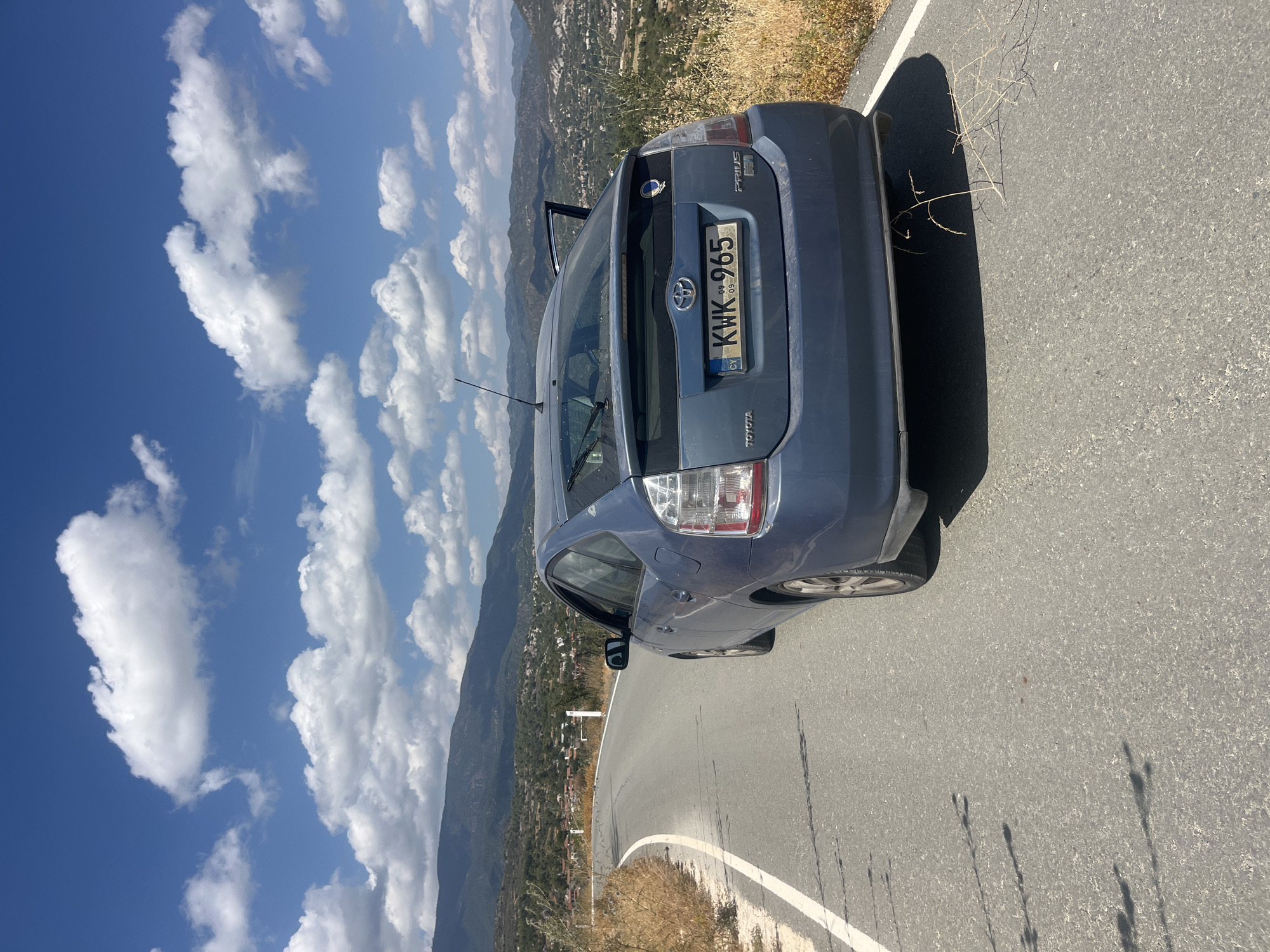 A blue Toyota Prius is parked on the side of a paved road that curves to the left. The car is viewed from the rear, with the driver's side door open. The license plate reads "KWK 965". The road is bordered by dry, yellow grass and shrubs on the right side. In the background, there are rolling hills covered in green vegetation and scattered buildings, indicative of a village. The sky is blue with scattered white clouds. The scene appears to be in a rural area, likely Silikou, Cyprus, given the context.