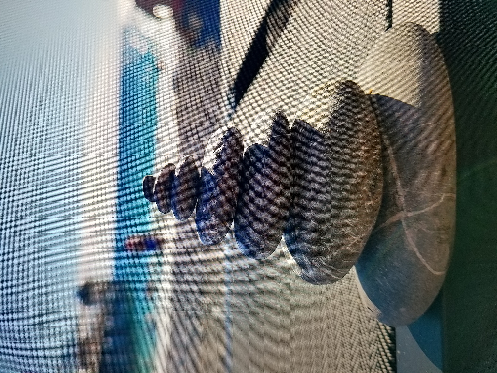 The image shows a stack of smooth, grey stones balanced on top of each other. In the background, a blurred beach scene with turquoise water and a pier is visible. There are a few out-of-focus figures in the distance. The stones are positioned on a textured surface, possibly a woven mat or fabric. The overall scene suggests a serene and calm atmosphere, evoking a sense of balance and nature. The image does not provide any specific details or context related to the city of Tiraspol, Moldova.