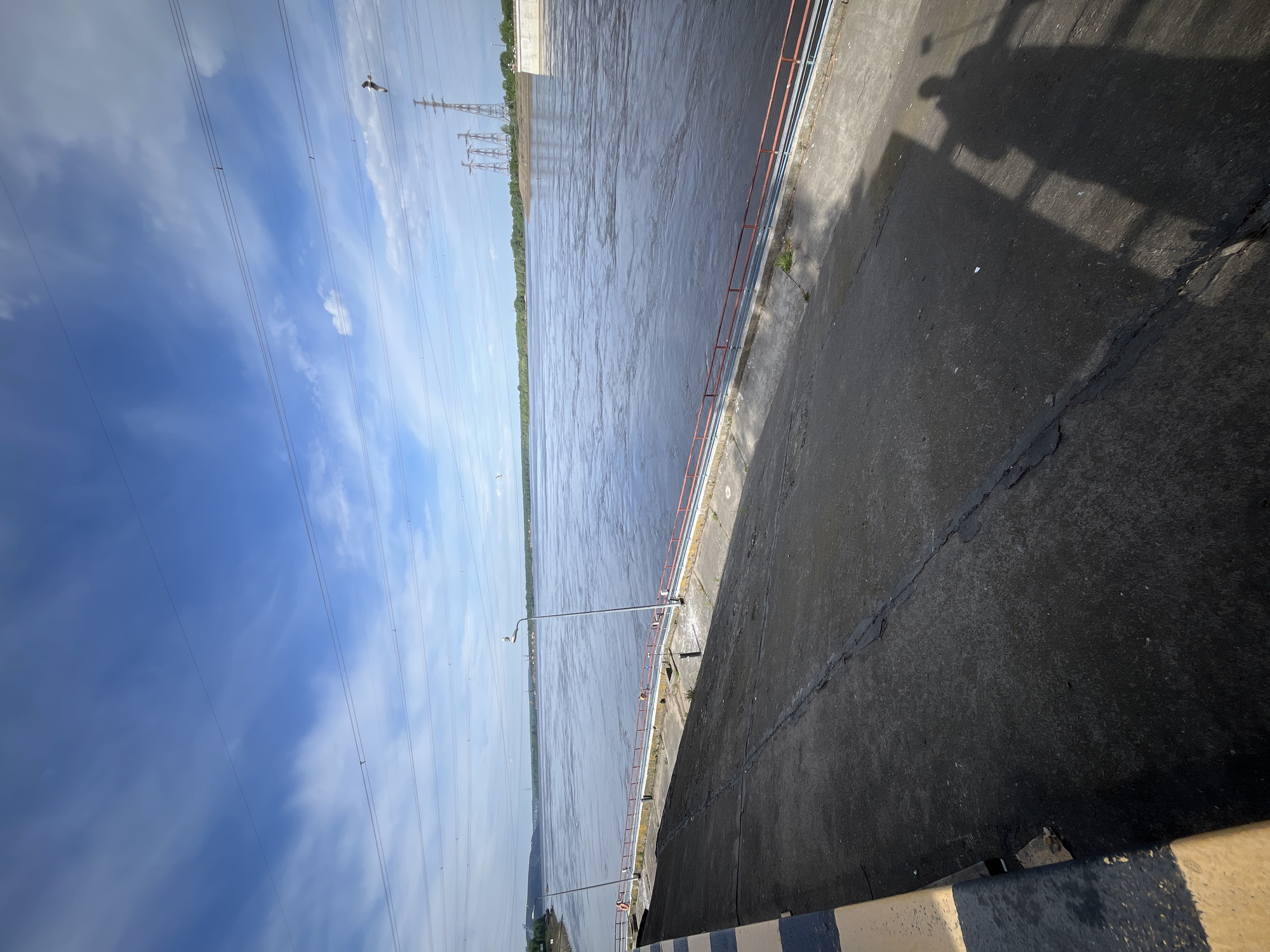 The image shows a concrete structure alongside a large body of water, possibly a reservoir or river, on a bright day. The concrete structure has a low metal railing along the edge. In the distance, there are electrical towers. The sky is blue with some scattered clouds. There is a shadow cast on the concrete. The image appears to be taken from a high vantage point looking down at an angle, resulting in the horizon being tilted. The location is likely Chaikovsky, Russia, given its proximity to large bodies of water and industrial infrastructure.
