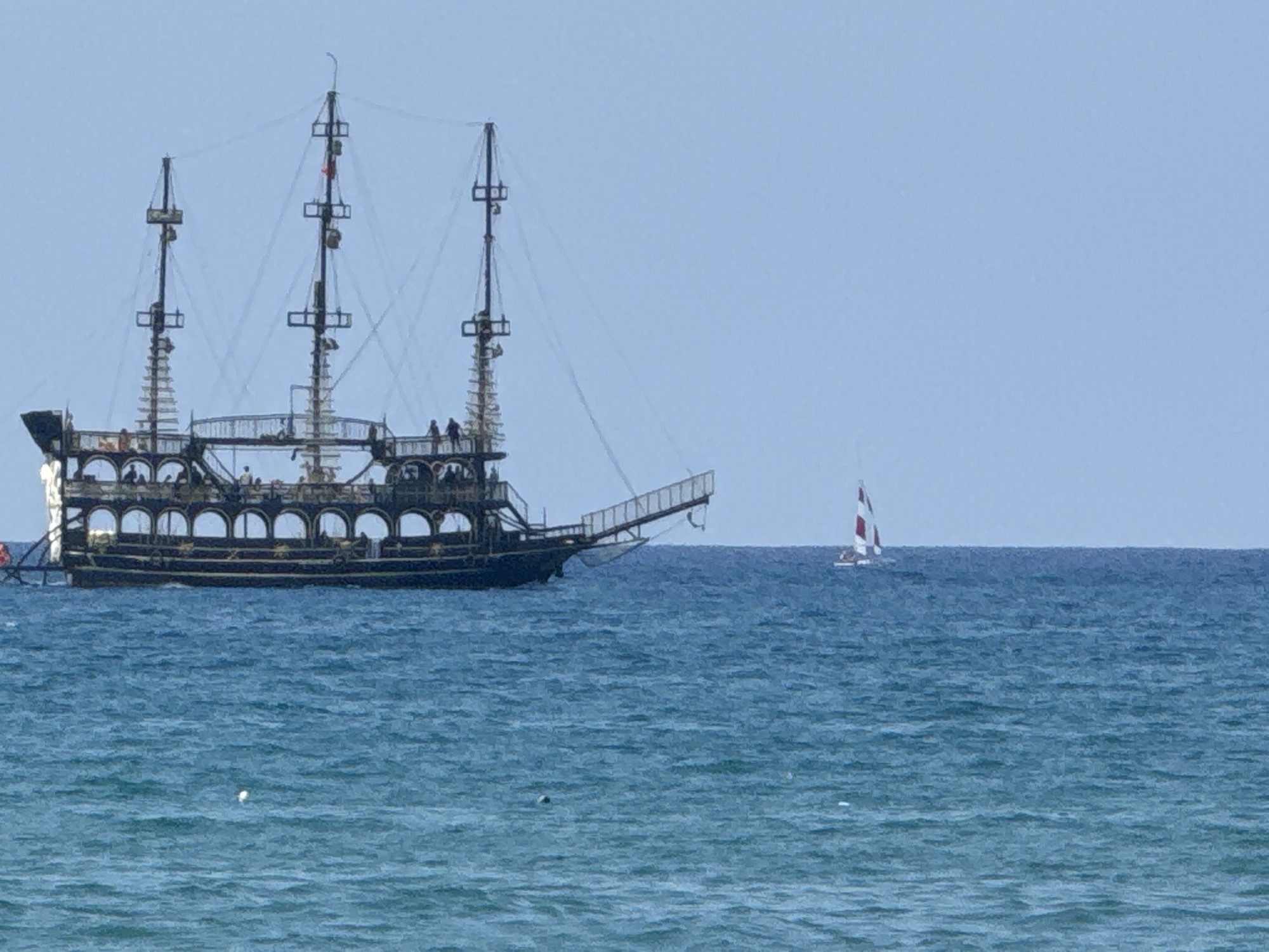 A large, black, pirate-style ship with three masts is sailing on the turquoise waters of the Mediterranean Sea, off the coast of Manavgat, Turkey. The ship has multiple decks and is adorned with arched windows. Several people are visible on the decks of the ship. To the right of the pirate ship, a smaller white sailboat with red trim is also sailing on the sea. The sky is a clear, pale blue. The water is slightly choppy, with small waves.