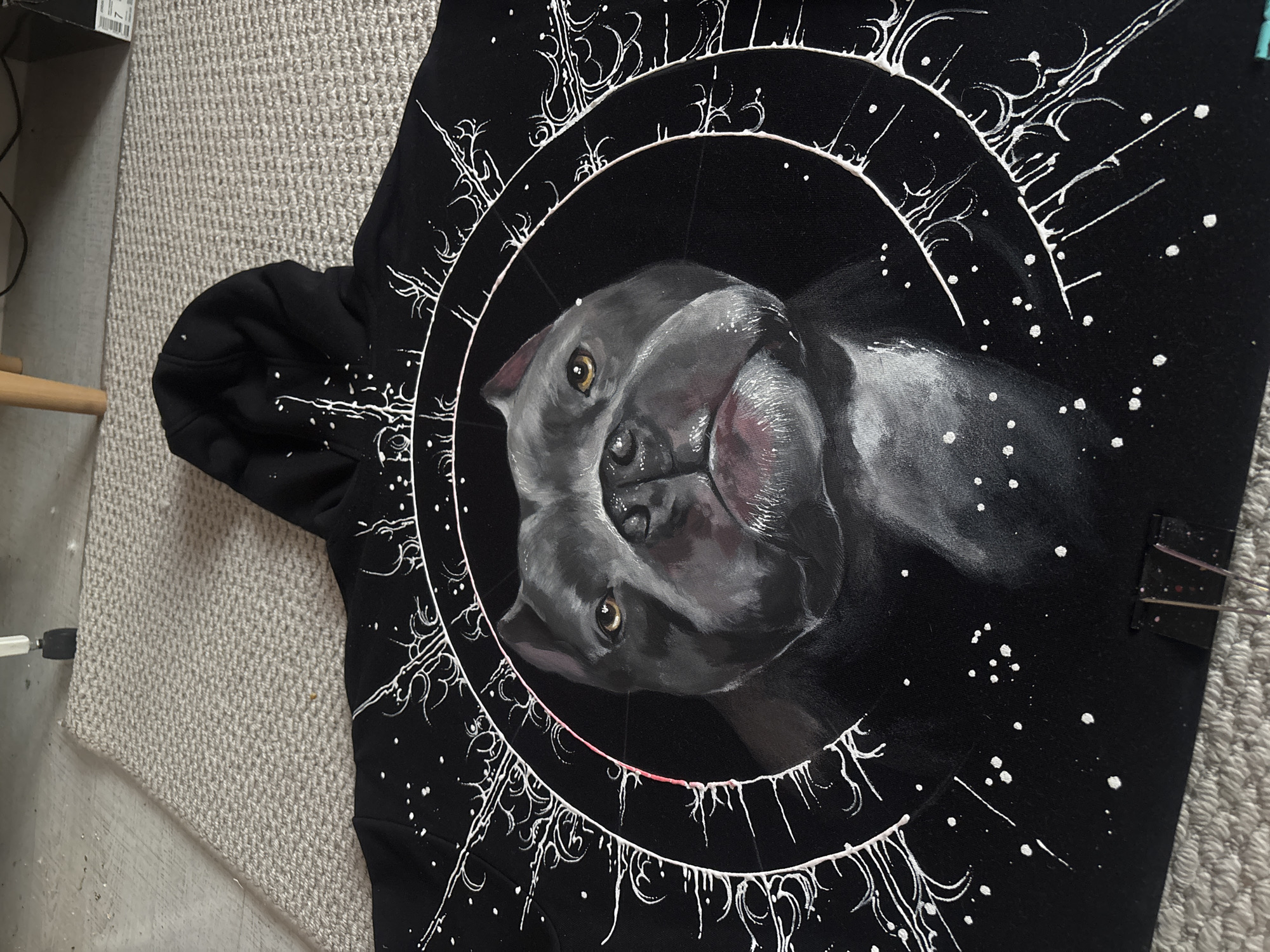 The image captures a black hoodie adorned with custom white paintwork, laid out on a light grey, textured carpet. The central feature of the hoodie is a detailed, realistic painting of a grey dog, resembling a Pit Bull or similar breed, with expressive golden-brown eyes and a prominent nose. The dog's head is depicted looking slightly upwards and to the right, encircled by a thick white circular frame.
Surrounding this central portrait are intricate, abstract designs executed in white paint. These patterns include multiple concentric rings, organic swirling motifs, and branching lines that emanate outwards, creating a celestial or mystical aura. Numerous small white splatters and dots are scattered across the black fabric, enhancing the cosmic effect. The white paint appears to have some texture, suggesting dimension.
In the background, the light grey carpet exhibits a distinct woven pattern. To the left of the frame, a segment of a light-colored wooden furniture leg is visible, along with a black box containing some faint, unreadable white text or labeling. Cables are also present near the box. On the far right edge of the hoodie, a black binder clip with a subtle speckled pattern is attached. The scene is set indoors, likely a room, with artificial lighting. The image showcases an artistic piece of clothing, implying it is either a display of artwork or an item for sale.
