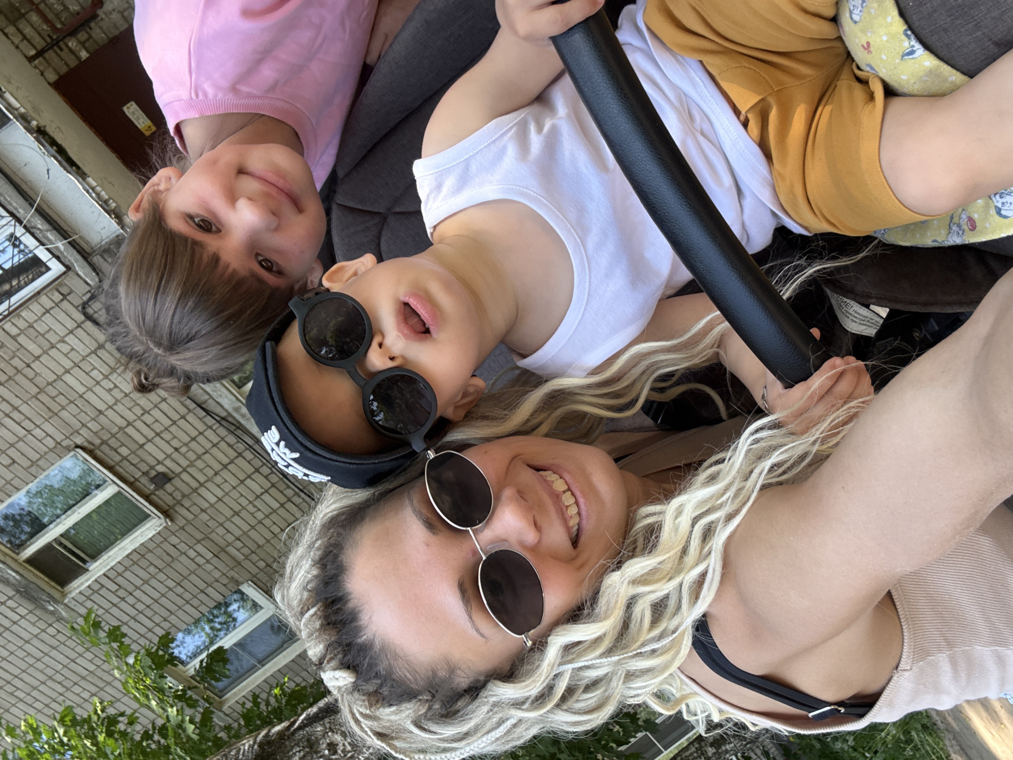 A selfie of a Caucasian woman with blond braided hair wearing round sunglasses and a beige top, a girl with brown hair wearing a pink top, and a young child wearing sunglasses, a black hat, and a white t-shirt. The child is sitting in a stroller with yellow shorts. The background features a brick building with windows and greenery. The location is Rusanovo, Russia.