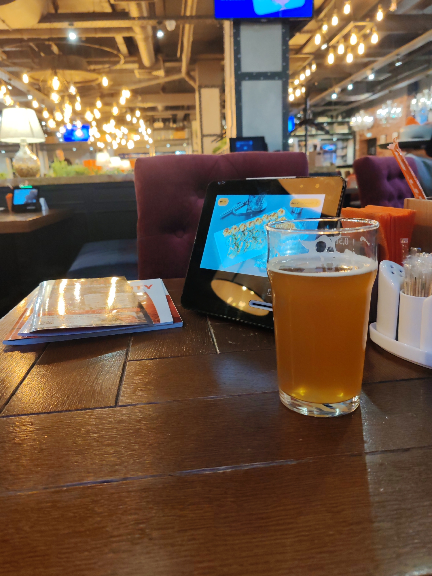This image provides a close-up view of a table in a restaurant or bar in Saint Petersburg, Russia, likely during the evening due to the warm, artificial lighting. In the foreground, a glass of amber-colored beer with a thin layer of foam sits on a dark, polished wooden table that displays a noticeable wood grain.
Beside the beer glass, a black tablet device is propped up, showing an image of sushi rolls on its screen. Partial Russian text is visible on the tablet, including "Как это..." (How is it/this...), suggesting an interactive menu or ordering system. To the left of the tablet, a stack of printed menus or magazines is neatly placed. To the right of the beer, a white condiment caddy holds salt and pepper shakers, along with a collection of toothpicks or stirrers.
In the midground, the back of a dark red or burgundy upholstered chair or booth provides a glimpse of the seating arrangement. The background reveals the establishment's interior design, characterized by an industrial-style ceiling with visible ducts and beams. Numerous Edison-style light bulbs hang, emitting a warm, inviting glow. A large television screen, displaying a bright blue image, is mounted on a structural pillar. Further back, more seating, ambient lighting, and a table lamp with a white shade contribute to a relaxed and comfortable dining or socializing environment.