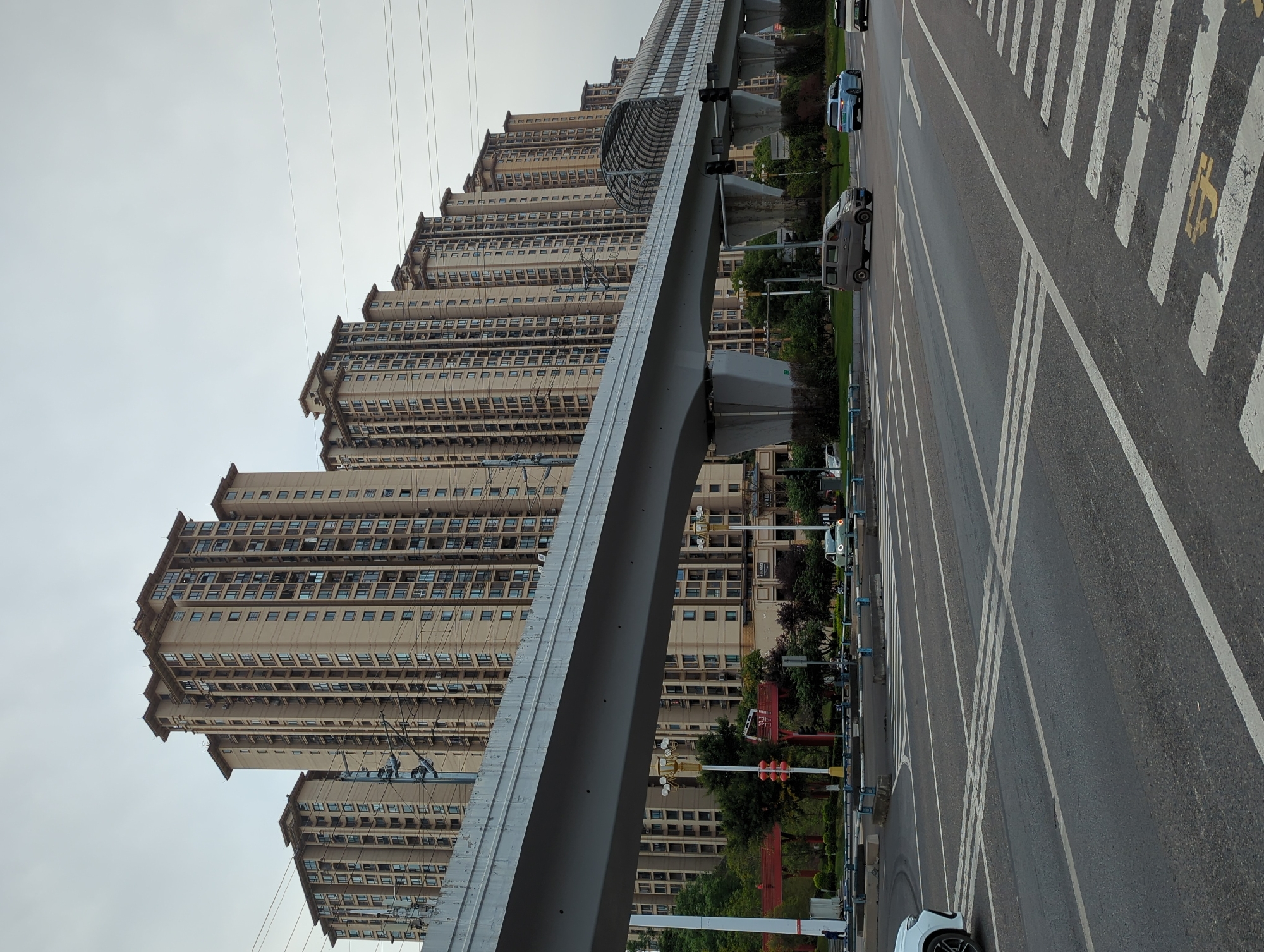 The image displays a modern urban landscape in Jiangjin, China, featuring towering residential buildings, an elevated transit system, and a multi-lane road under an overcast sky.
On the left, multiple high-rise apartment buildings with a stepped architectural design dominate the background. These buildings are uniformly colored in a light beige or cream hue, featuring numerous windows and what appear to be small balconies or ledges on each floor. Overhead, several utility wires crisscross the sky.
In the mid-ground, a prominent, sleek, gray elevated track, likely for a monorail or light rail, runs parallel to the buildings. This structure is supported by robust concrete pillars and includes a section with a distinctive translucent, arched covering.
To the right, a wide asphalt road with at least three lanes is visible. It features clear white road markings, including solid lines, dashed lines, and multiple pedestrian crosswalks (zebra crossings). A yellow Chinese character "停" (tíng), meaning "stop," is painted on the road within one of the crosswalk areas. Several vehicles are present on the road: a dark gray minivan, a light blue car, and the front of a white car in the immediate foreground. Another light-colored SUV is further down the road.
The roadside is lined with trees and green shrubbery, along with streetlights, traffic signals, and blue-colored protective barriers. The general scene suggests a busy city thoroughfare, possibly a residential or commercial area, with no individuals explicitly visible. The weather appears to be cloudy, providing soft, diffused lighting.