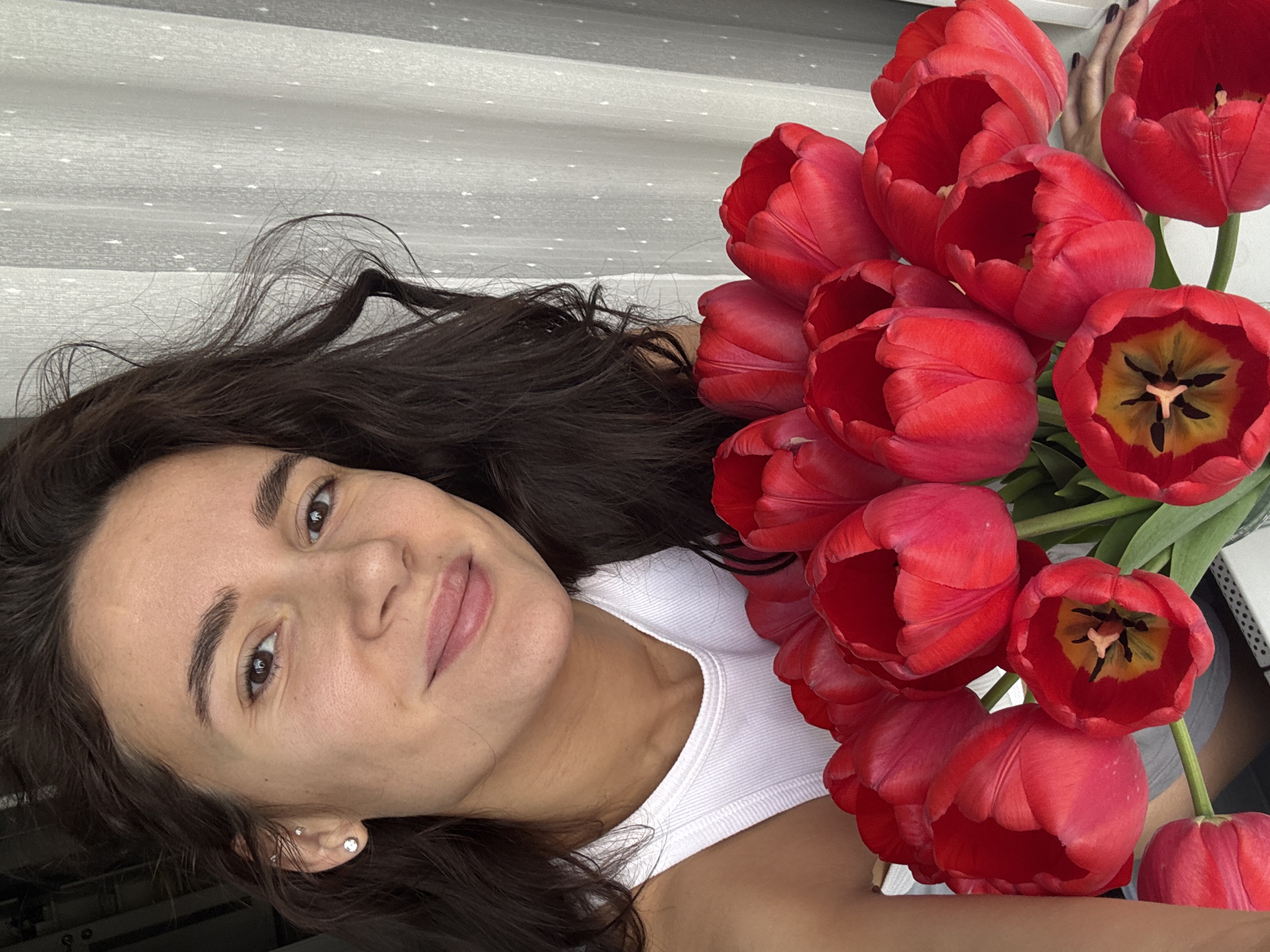A young woman with dark hair and fair skin is shown in a selfie-style photograph, holding a bouquet of red tulips. The woman is wearing a white tank top. She is smiling and looking at the camera. The background appears to be an interior space with a textured or patterned surface. It is impossible to confirm the location as the city of Chaikovsky, Russia with the given image.