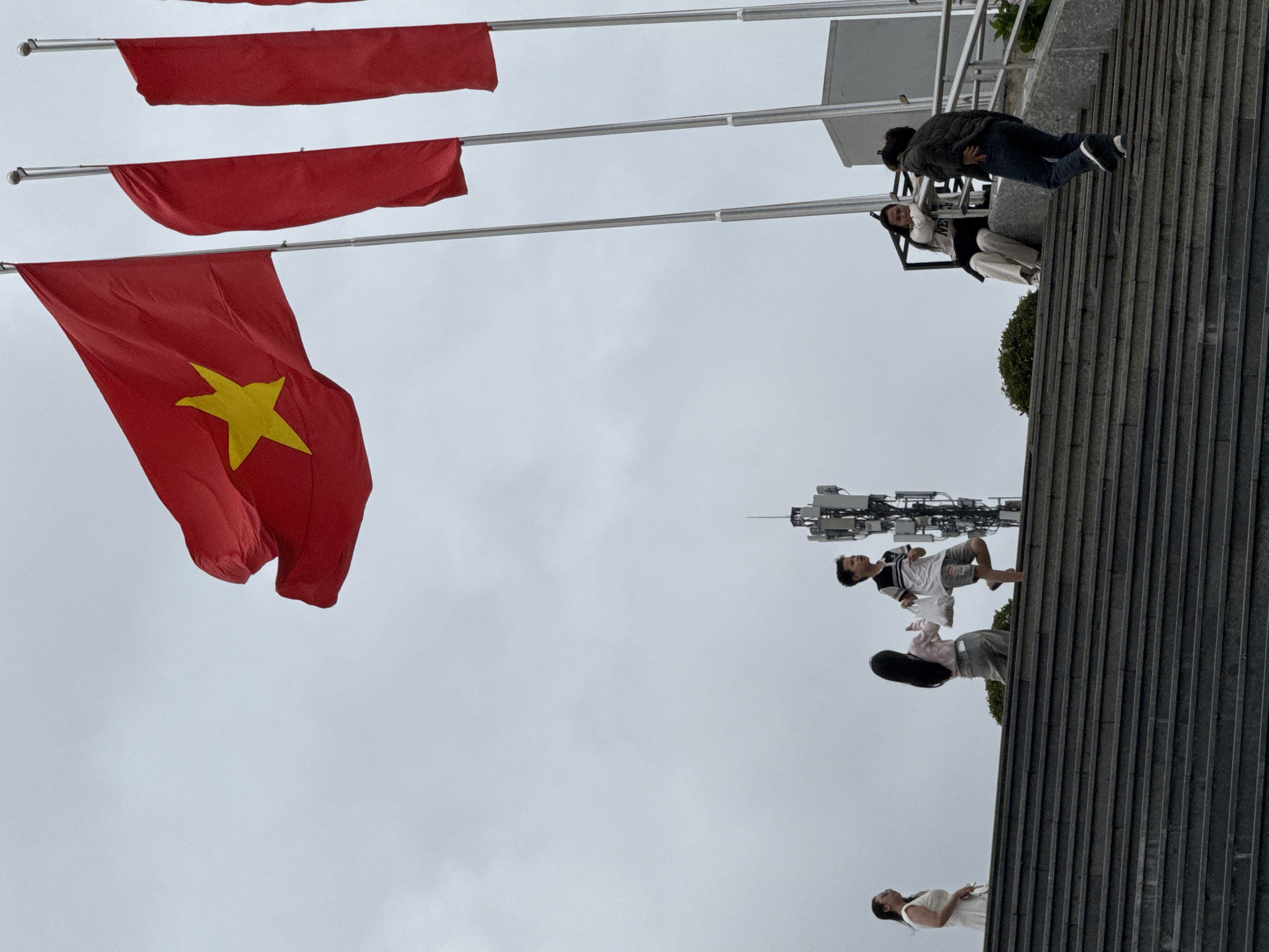 The image captures a wide shot of a grand staircase leading upwards, possibly towards a memorial or monument, under a cloudy sky. Dominating the upper portion of the frame are large Vietnamese flags, a prominent red flag with a yellow star is waving prominently, accompanied by two other red flags on tall poles. On the staircase, several people are visible. On the far left, a woman in a white dress is looking towards the left. Moving up the stairs, a young girl with long black hair is holding two white plastic bags. To her right, a young boy in a striped t-shirt and shorts is walking, also carrying two white plastic bags. Further up and to the right, a woman is seated on a bench, wearing a light-colored sweater and pants, and a man in a dark jacket and jeans is walking up the stairs. In the background, a tall cell tower rises against the overcast sky, suggesting an urban or semi-urban setting. The overall atmosphere is somber due to the cloudy weather, but the presence of the flags and people suggests a place of significance. This scene is consistent with locations like the Da Lat Flower Garden or other public spaces in Da Lat, Vietnam, that feature prominent flagpoles and open areas for gatherings.