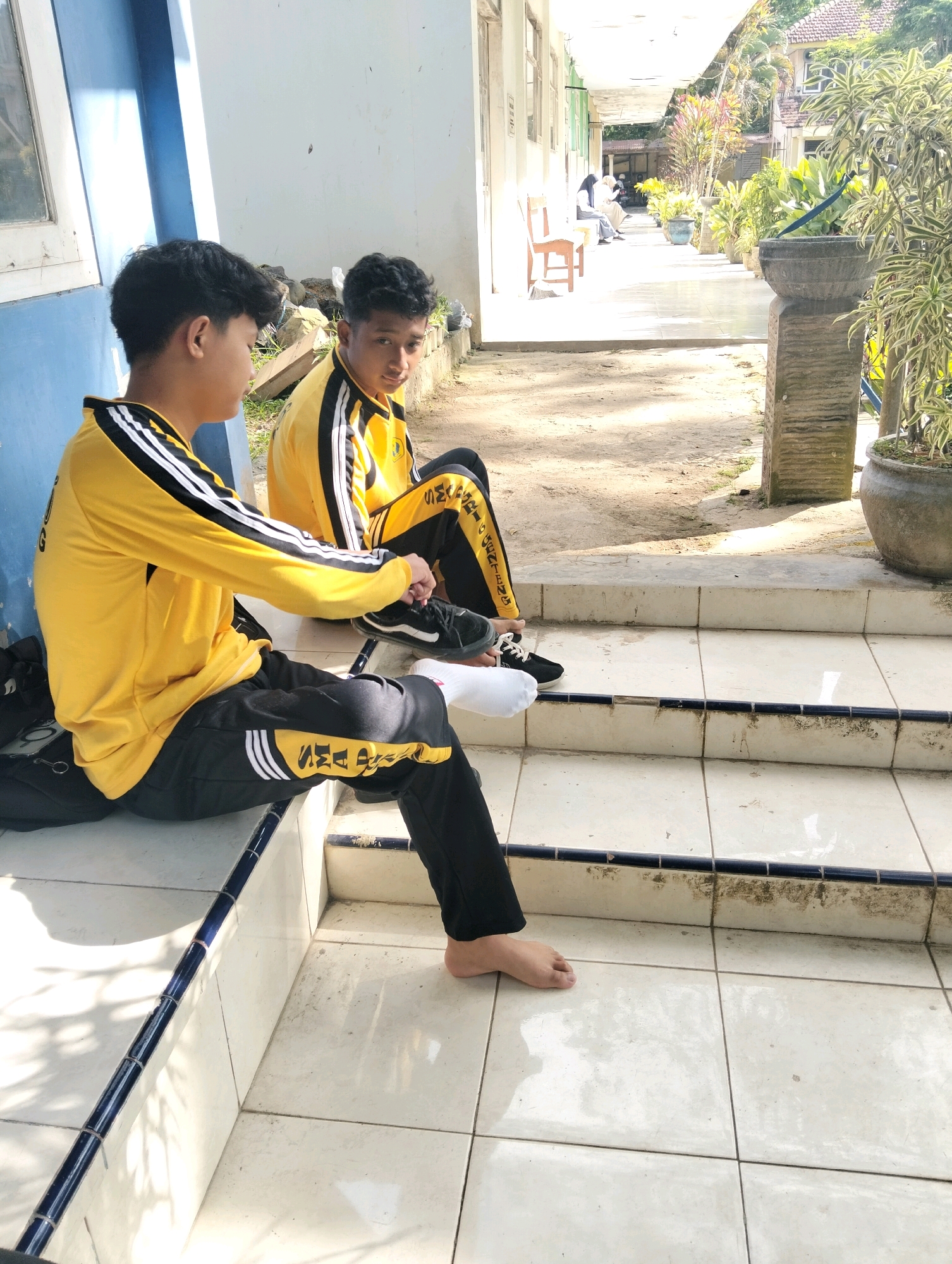 Two teenage boys are sitting on the tiled steps of a building, possibly a school or community center, in Genteng, Indonesia. The boy on the left, with dark hair, is wearing a yellow long-sleeved shirt with black and white stripes on the sleeves and black pants with yellow and white stripes. He is tying his black and white sneakers. The boy on the right, also with dark hair, is looking towards the camera and is wearing a similar yellow shirt and black pants. The background shows a walkway with potted plants and other buildings. The lighting suggests it is daytime, and there is a shadow cast on the steps, indicating sunlight. The environment appears to be outdoors, with a dusty ground and some greenery.