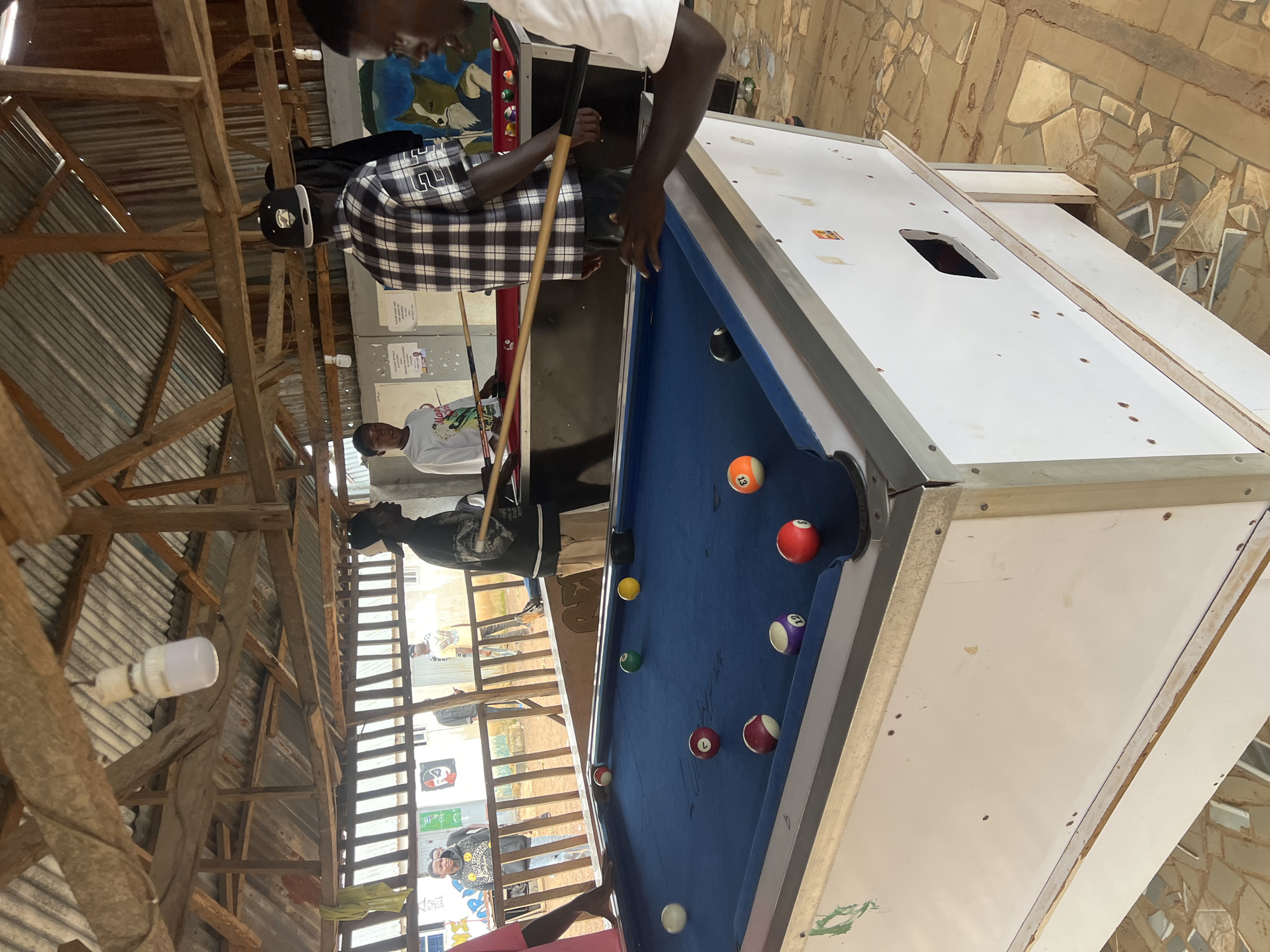 In Durumi, Nigeria, a group of young men are engaged in a game of pool. The setting appears to be an outdoor, open-air establishment with a rustic, unfinished wooden roof supported by beams. A fluorescent light bulb hangs precariously from the roof. The pool table itself is a standard size with a blue felt surface and white bumper. Several billiard balls are scattered across the table, including a red 3-ball, a purple 12-ball, a red 5-ball, and an orange 13-ball. One man in a black and white checkered shirt and a black baseball cap is poised to strike a ball with a pool cue. Another man in a black t-shirt and light-colored pants stands nearby, observing the game. In the background, other individuals are visible, some standing and others potentially involved in the game. The walls behind the pool table are decorated with colorful murals and posters. The ground is paved with irregular stones.