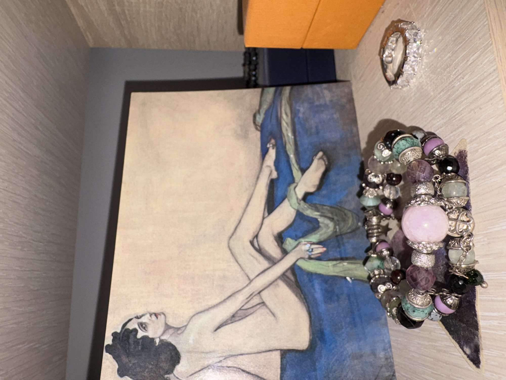 A photograph shows a still life of objects arranged on a light wood-grain surface with a blue-gray interior backdrop. The items consist of a framed print featuring a reclining nude woman with black hair against a tan background and blue accents, an orange rectangular box, a silver ring with multiple white stones, and a bracelet with a variety of colorful beads and charms. The scene is likely indoors, given the context of the objects. There is no definitive information within the image to confirm its location in Izhevsk, Russia.