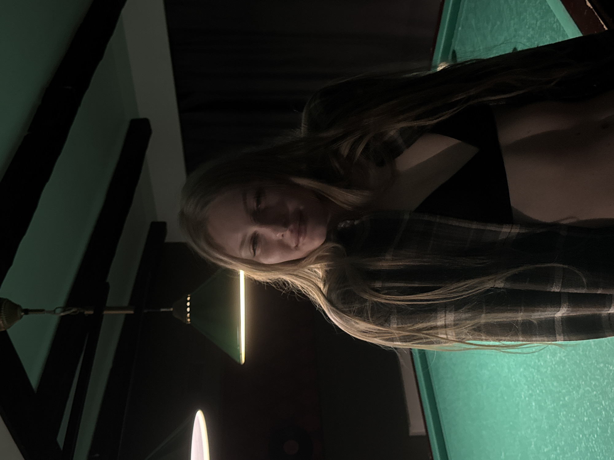 The image depicts a female subject posing indoors, likely within a billiards establishment in Perm, Russia. The environment is dimly lit. The subject, a woman with long, light brown hair, is positioned centrally, facing forward with a slight smile. She is wearing an open plaid shirt over a black bra, exposing her midriff. Behind her, a green-felted billiards table is visible. Overhead, two illuminated lamps are present: one with a white circular shade on the left, and another with a green conical shade in the center. A third, decorative, unlit fixture hangs further back. The background consists of dark walls or drapes and a dark ceiling with visible beams. No specific landmarks or unique identifiers within Perm are discernible from the image.