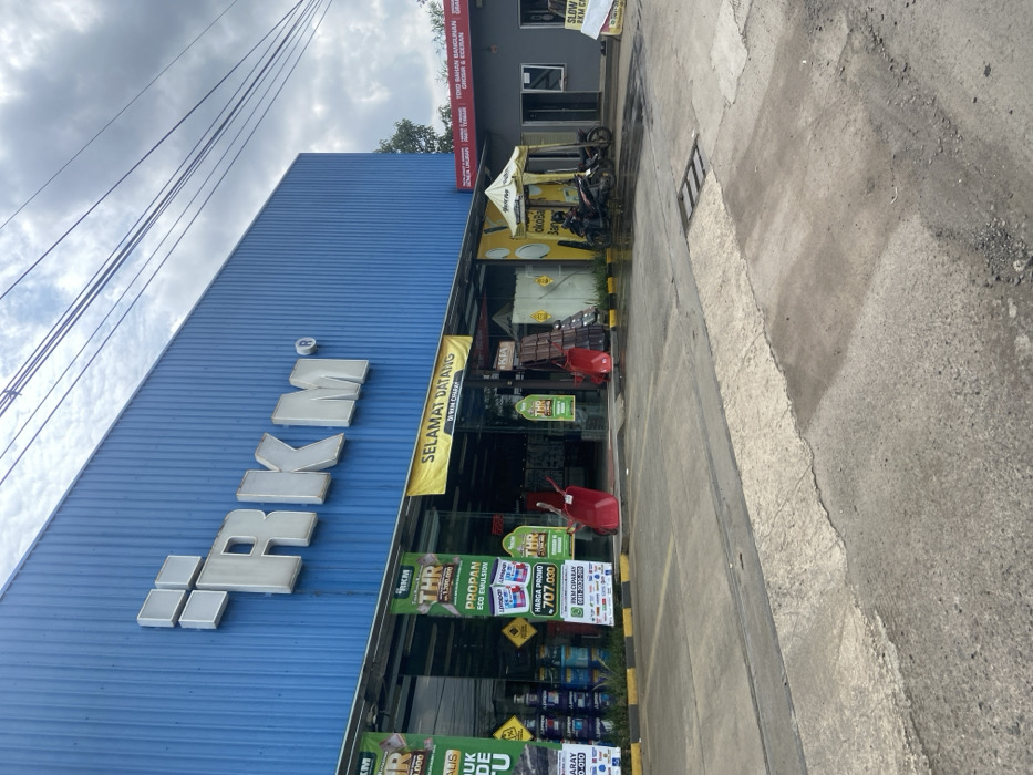 The scene is in front of an RKM building located in Ciparay, Indonesia, at latitude -7.029 and longitude 107.697. The building is a large, blue structure with the word "RKM" in white lettering on the front. There are several signs and posters displayed in front of the building, advertising various products and services, including "THR," "Propan Eco Emulsion," and "Harga Promo." The posters are in Indonesian. Two red wheelbarrows are leaning against the front of the building. There is a small, yellow-and-white tent set up in front of the building. A person is standing inside the tent. A motorcycle is parked in front of the tent, and there is a second motorcycle parked on the sidewalk. The sidewalk is paved with concrete and is slightly wet. There is a small drainage ditch in the middle of the sidewalk. The sky is overcast with a few clouds. There is a sign on the right side of the image that reads "TOKO BANGUNAN, BANGUNAN, GROSIR & ECERAN."