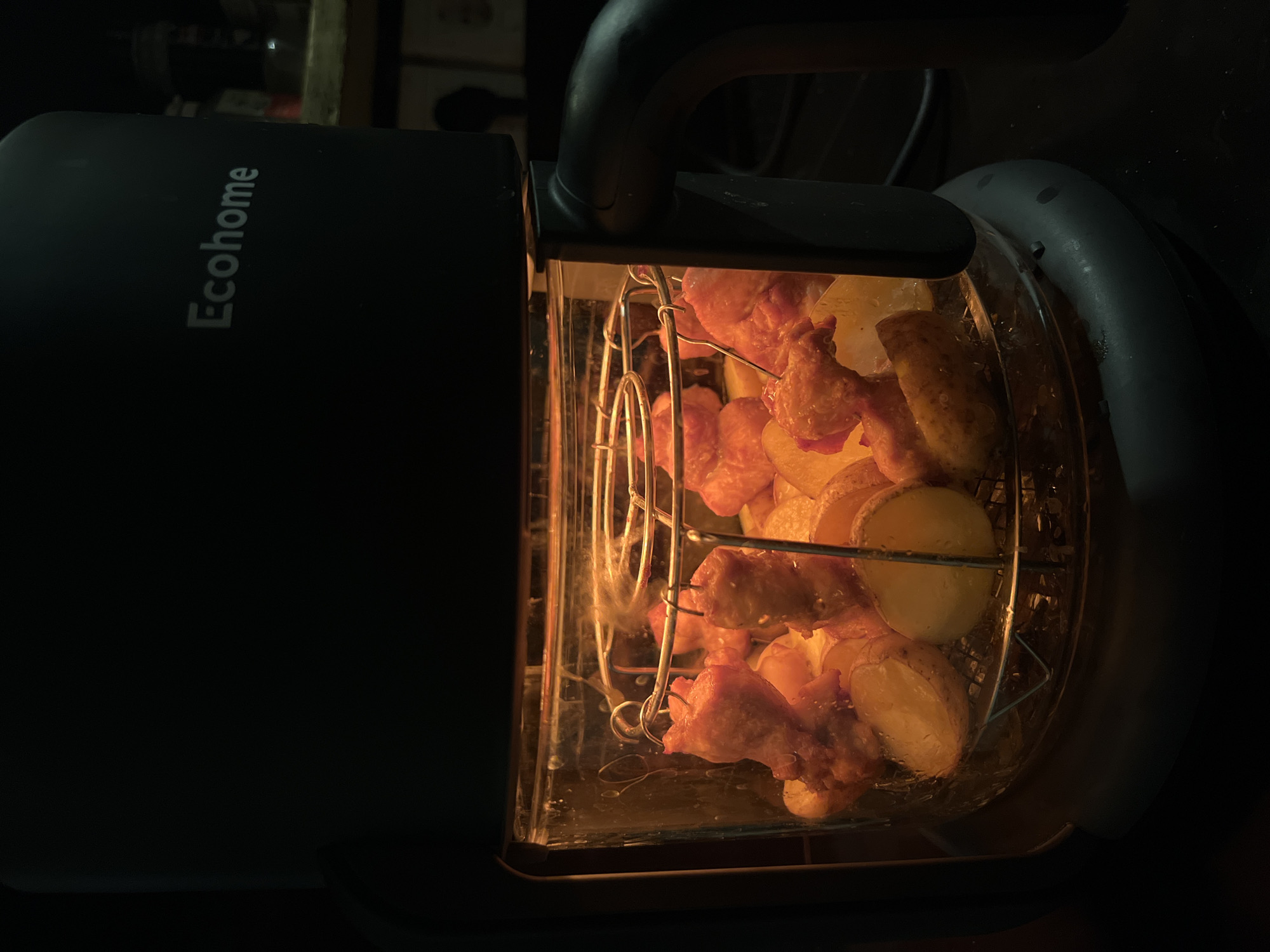 The image depicts food being cooked in an Ecohome brand air fryer. The food appears to be chicken wings and sliced potatoes, arranged in a wire rack inside the appliance. The air fryer is emitting light from within, suggesting it is in operation. The background is dark and indistinct, but elements of a room are suggested. There is no evidence to indicate the location is Sukawati, Indonesia.