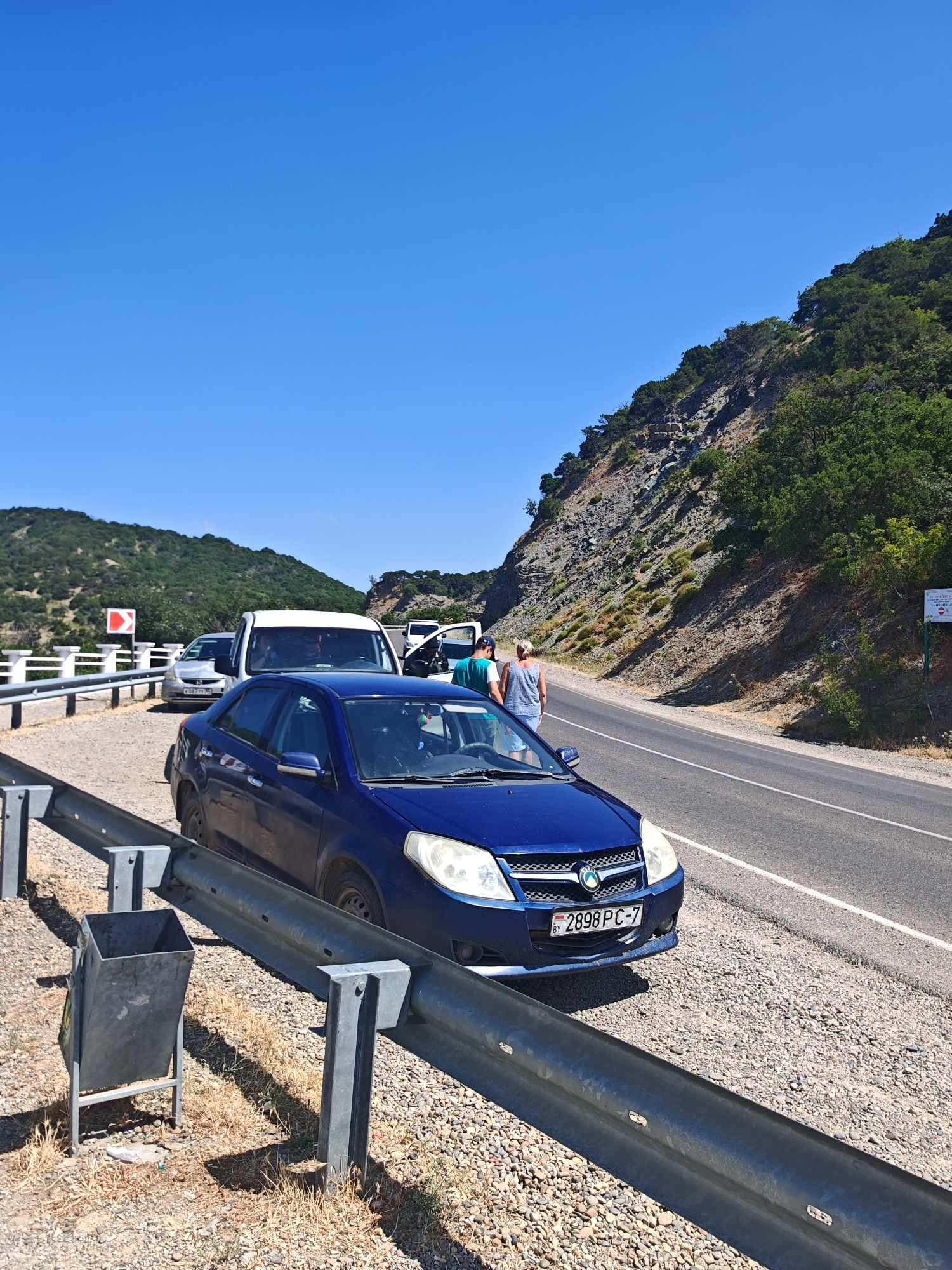 The image shows a traffic jam on a paved road in Sukko, Russia, near a steep, vegetated mountainside. The road is lined with a metal guardrail, and a gray trash can is visible on the shoulder. In the foreground, a blue sedan is stopped on the road. Behind it, a white van and a silver car are also stopped. Several people are standing on the side of the road, some near the vehicles, and others appear to be walking. The sky is clear and blue.