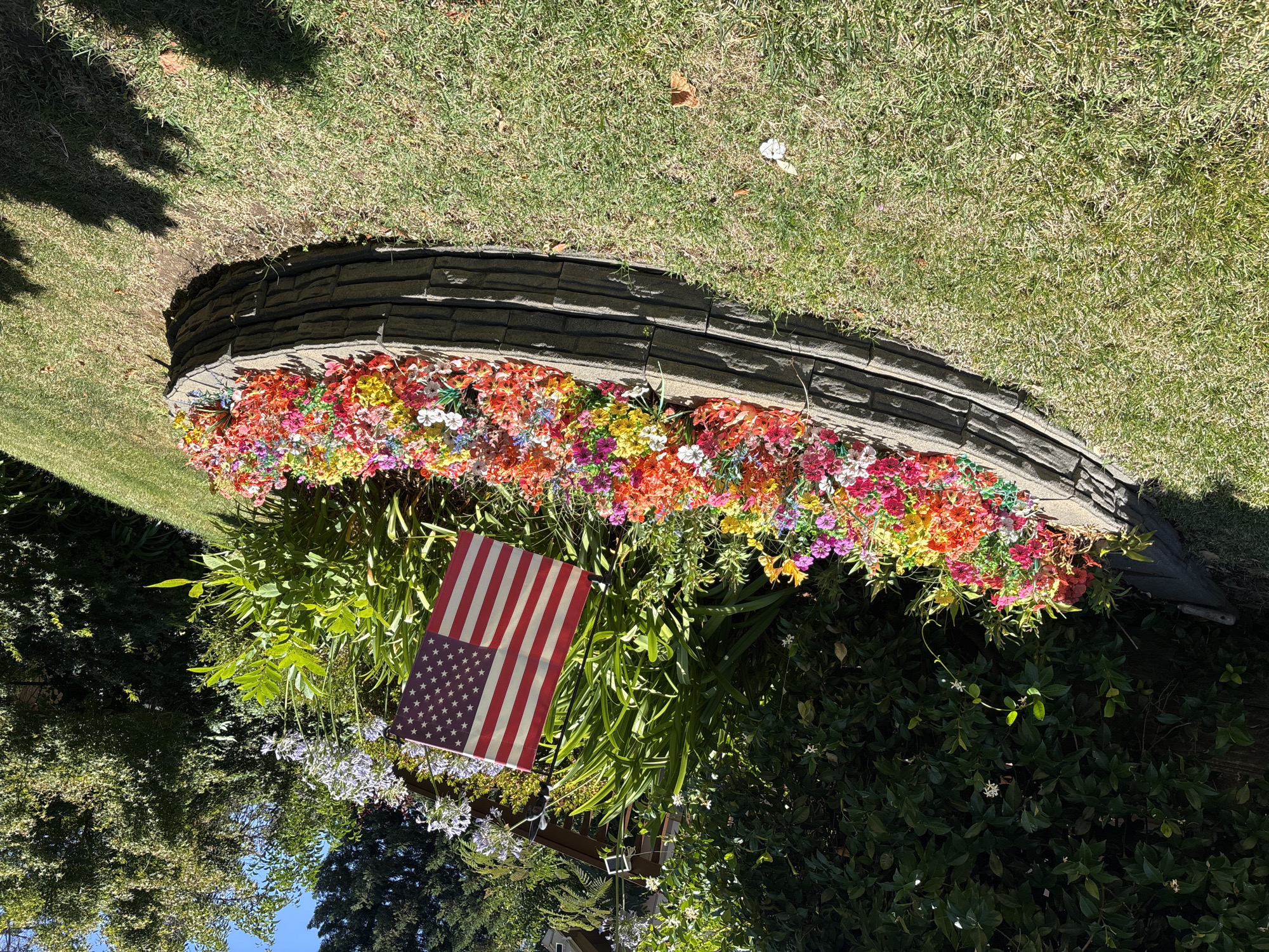 In the city of Orangevale, United States, a raised flower bed, constructed from stacked gray stones, is filled with a vibrant assortment of artificial flowers in various shades of red, orange, yellow, pink, and purple. An American flag is positioned in the center of the flower bed, leaning slightly to the right. The flower bed is situated on a grassy lawn, which appears somewhat dry in patches. Behind the flower bed, lush green foliage and trees form a backdrop. In the upper right portion of the image, part of a residential structure with a wooden deck is visible amongst the greenery. The scene is illuminated by bright sunlight, casting distinct shadows on the grass.