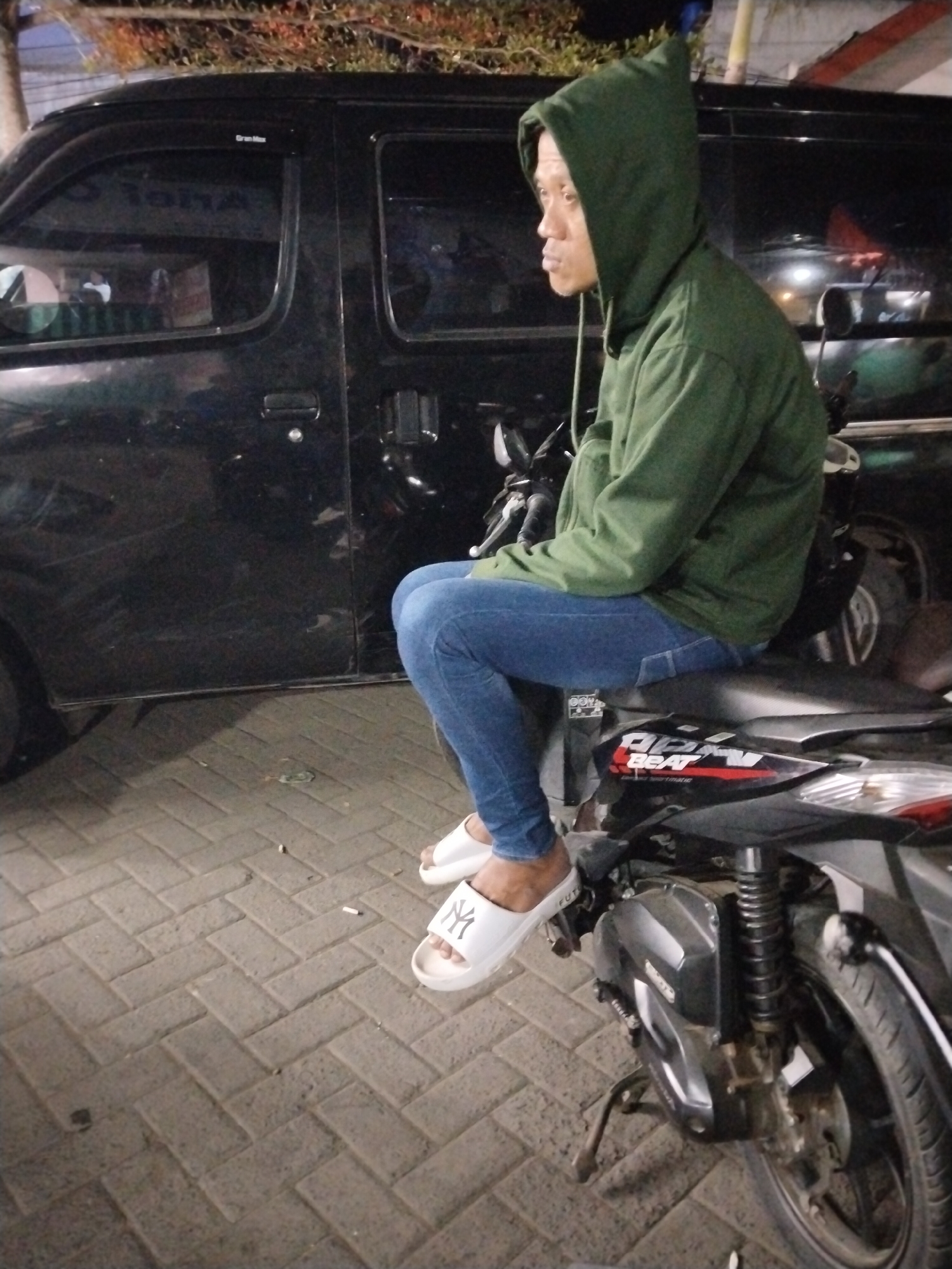 A person is sitting on a motorcycle in front of a black van. The person is wearing a green hooded sweatshirt and blue jeans. They are also wearing white slides with a New York Yankees logo on them. The motorcycle is black and has red and white decals on the side. The van is black and appears to be a passenger van. The scene is set on a brick-paved area, likely a street or parking lot in Tasikmadu, Indonesia. The lighting suggests it is nighttime or dusk. There are some trees or foliage visible in the background.