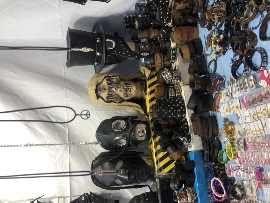 A sideways view displays a tabletop assortment. Black steampunk-style masks and headwear are displayed, including a top hat with goggles and a plague doctor mask. A scarecrow-like head with a burlap hood is also visible. Adjacent to these items are various leather bracelets and wristbands, some with studded embellishments. Further along, a collection of jewelry and accessories with plastic packaging and price tags can be observed. A peace sign necklace hangs nearby.