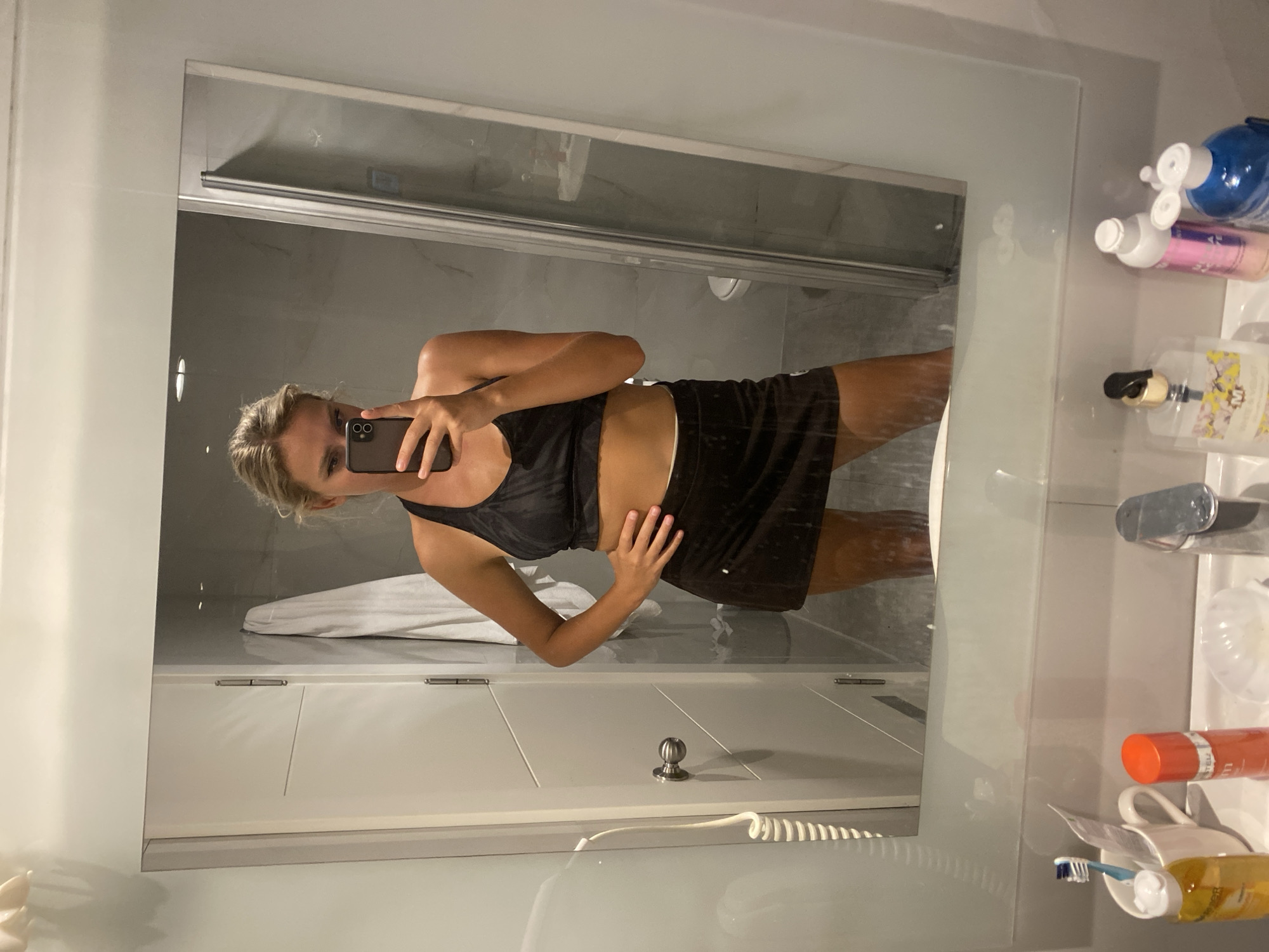 A woman stands in a bathroom, taking a selfie in a mirror. She is wearing a black sports bra and a dark brown skirt. Her hair is blonde and pulled back. Her left hand is on her hip, and she is holding a smartphone in her right hand, angled to capture the reflection. The bathroom appears to have white tiled walls and a glass shower enclosure. In the foreground, a sink is visible with various toiletries, including a toothbrush, toothpaste, and bottles of liquid. The overall impression is of a private space, likely a home bathroom. There is no indication of the city of Aksu, Turkey.