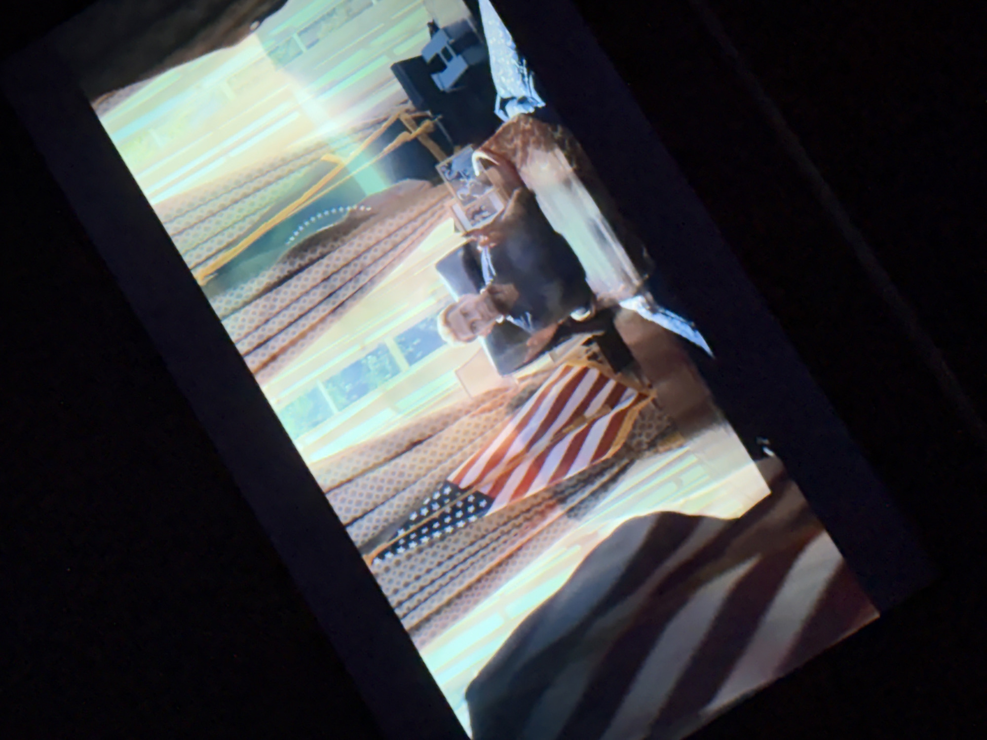 The image shows a dimly lit room with a screen displaying a scene. On the screen, a man with blond hair is sitting in a chair, holding a framed picture. An American flag is visible in the foreground of the screen. The background of the screen shows curtains and a window with sunlight streaming through. The room itself is dark, with the screen being the primary source of light. There is no information to indicate that this scene is located in Dnipro, Ukraine.