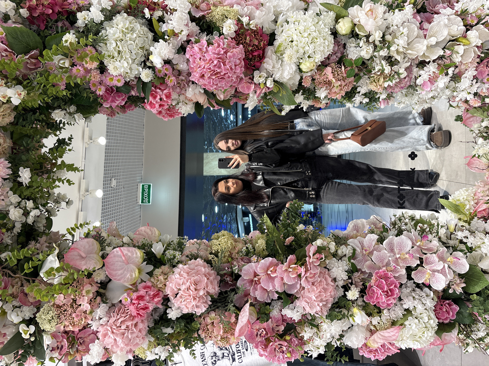 Two women are taking a selfie in front of a flower-covered mirror. The mirror reflects their images, showing one woman in a black leather jacket and black jeans, and the other in a black leather jacket and light-wash jeans. The flower arrangement surrounding the mirror consists of pink, white, and green flowers. The reflection in the mirror shows a shop interior with fluorescent lighting and an exit sign in Russian (выход). The location is the Fashion City shopping mall in Yekaterinburg, Russia, as identified by writing on a T-shirt that is also reflected in the mirror.