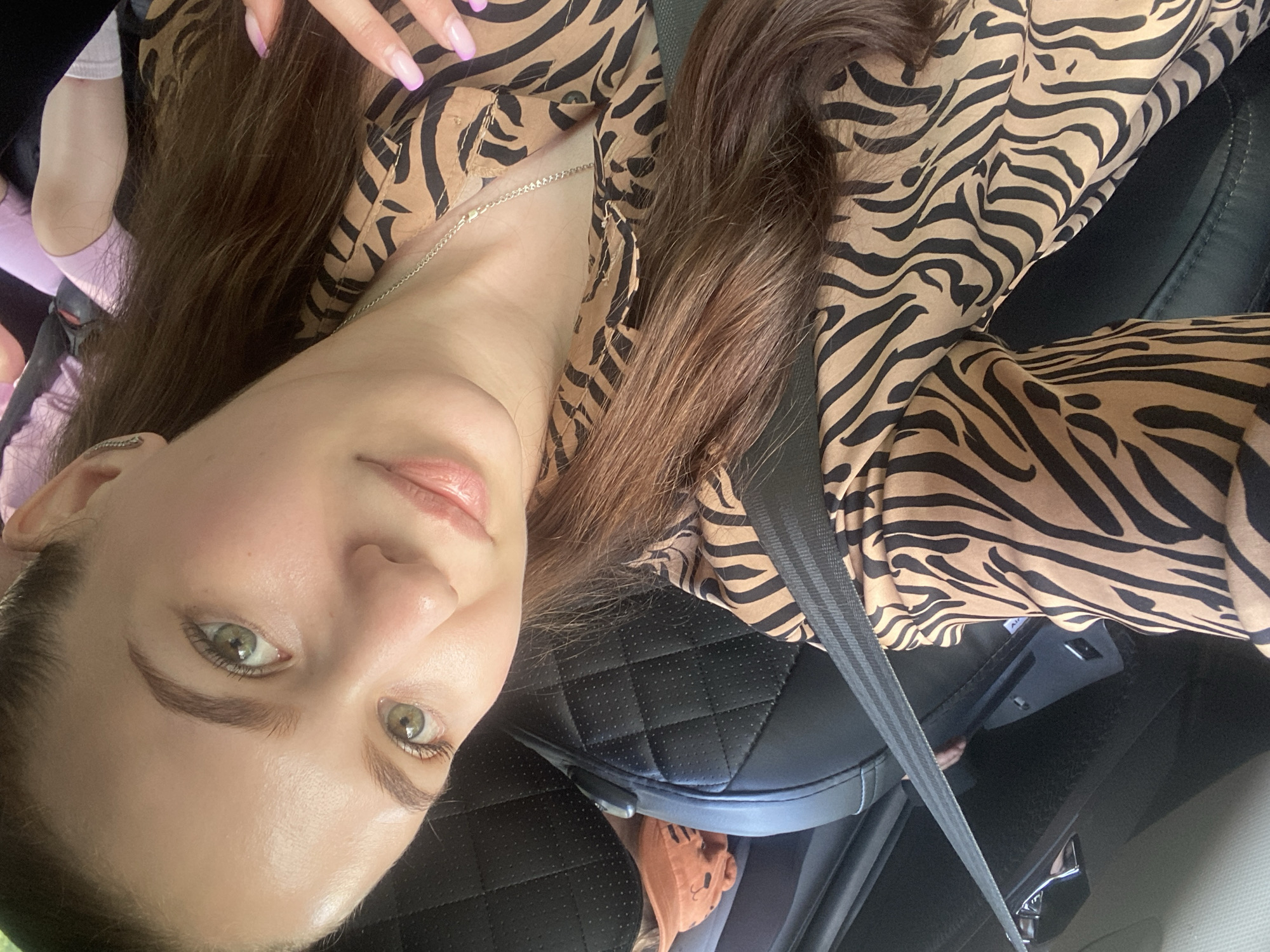 A woman is shown in a close-up selfie, wearing a seatbelt and a tan and black zebra-striped shirt. Her fingernails are painted a light purple. She has brown hair and green eyes. The background appears to be the interior of a car, specifically the passenger seat, with black leather upholstery. The location in the city of Chaikovsky, Russia, cannot be determined from the image.