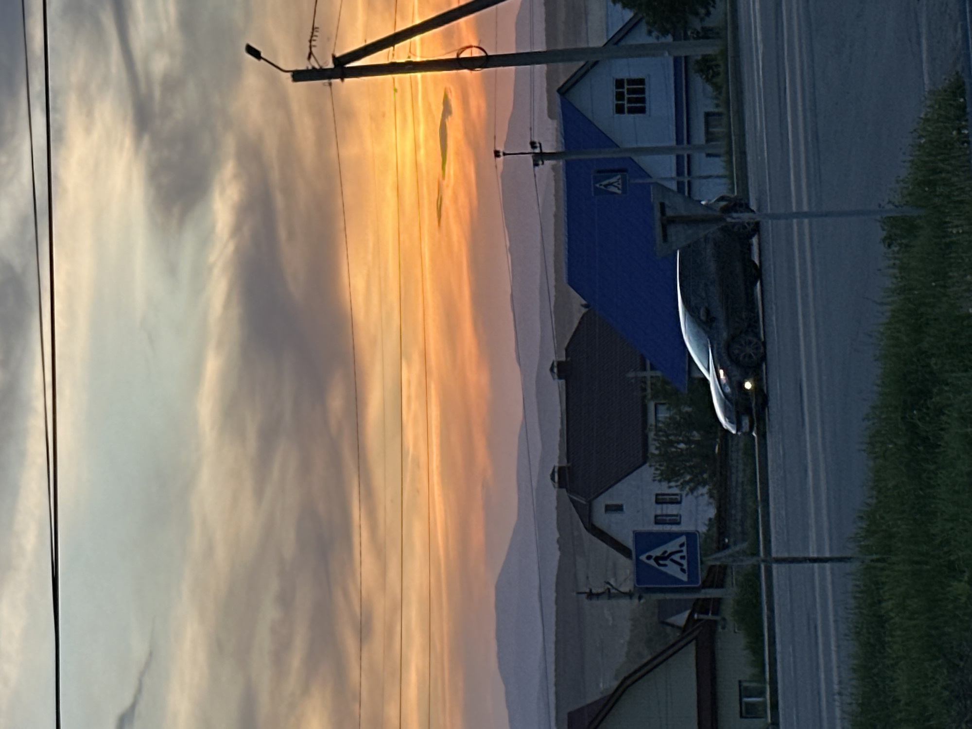 The image depicts a street scene at sunset in an unknown location. A dark-colored car is driving on a paved road. Adjacent to the road is a grassy area. A pedestrian crossing sign is visible near a building with a blue roof. In the background, there are mountains and a sky with orange and gray clouds, indicative of sunset. Electrical wires and a street light are also present.