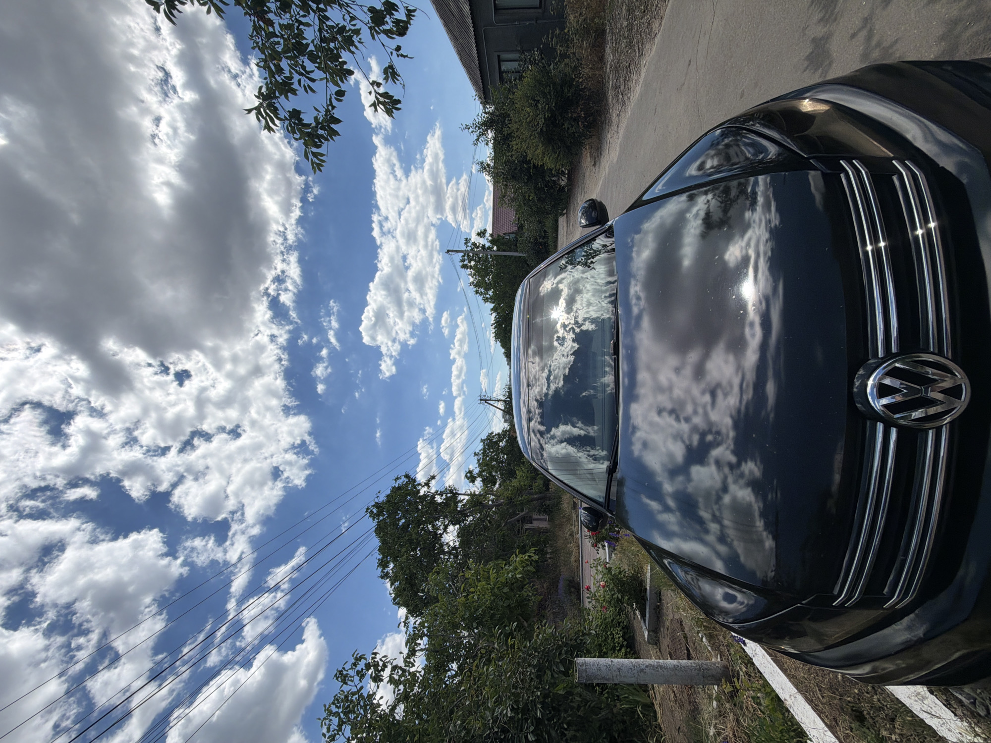 The image is an outdoor, eye-level shot of a black Volkswagen car parked on a dirt road with some vegetation and buildings in the background. The car's hood and grille are the primary focus, with the sky and clouds reflected on the polished surface. The sky is a bright blue with numerous white, puffy cumulus clouds. Trees and foliage line the road and surround the buildings. The scene appears to be set during the daytime, with the sun casting bright reflections on the car, suggesting a sunny or partly cloudy day. The overall mood is calm and perhaps a bit serene, with the bright sky and reflections creating a pleasant visual. There is no visible text in the image.